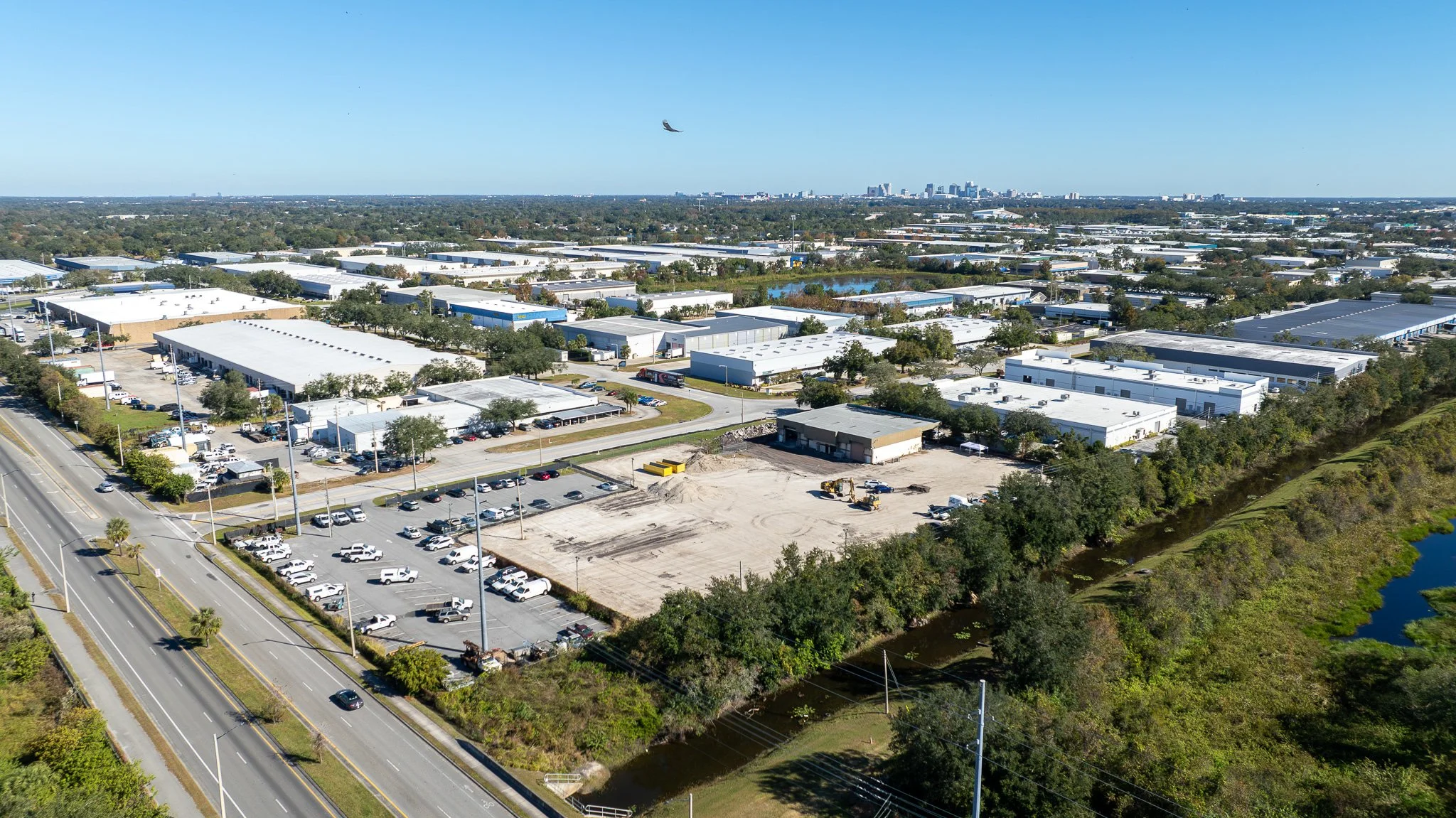 Aerial view of an industrial park with several large warehouse buildings, parking lots, and a construction area with excavators, next to a road and a small river, with a city skyline in the distance under a clear blue sky.