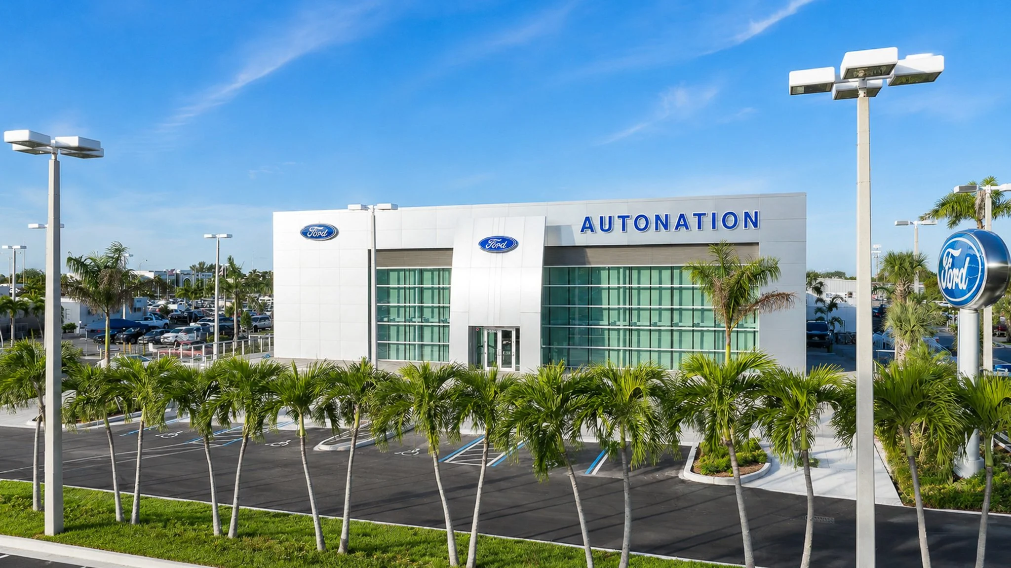 Exterior of a Ford dealership with a sign that says 'AUTONATION', surrounded by palm trees, with parking lot and multiple light poles