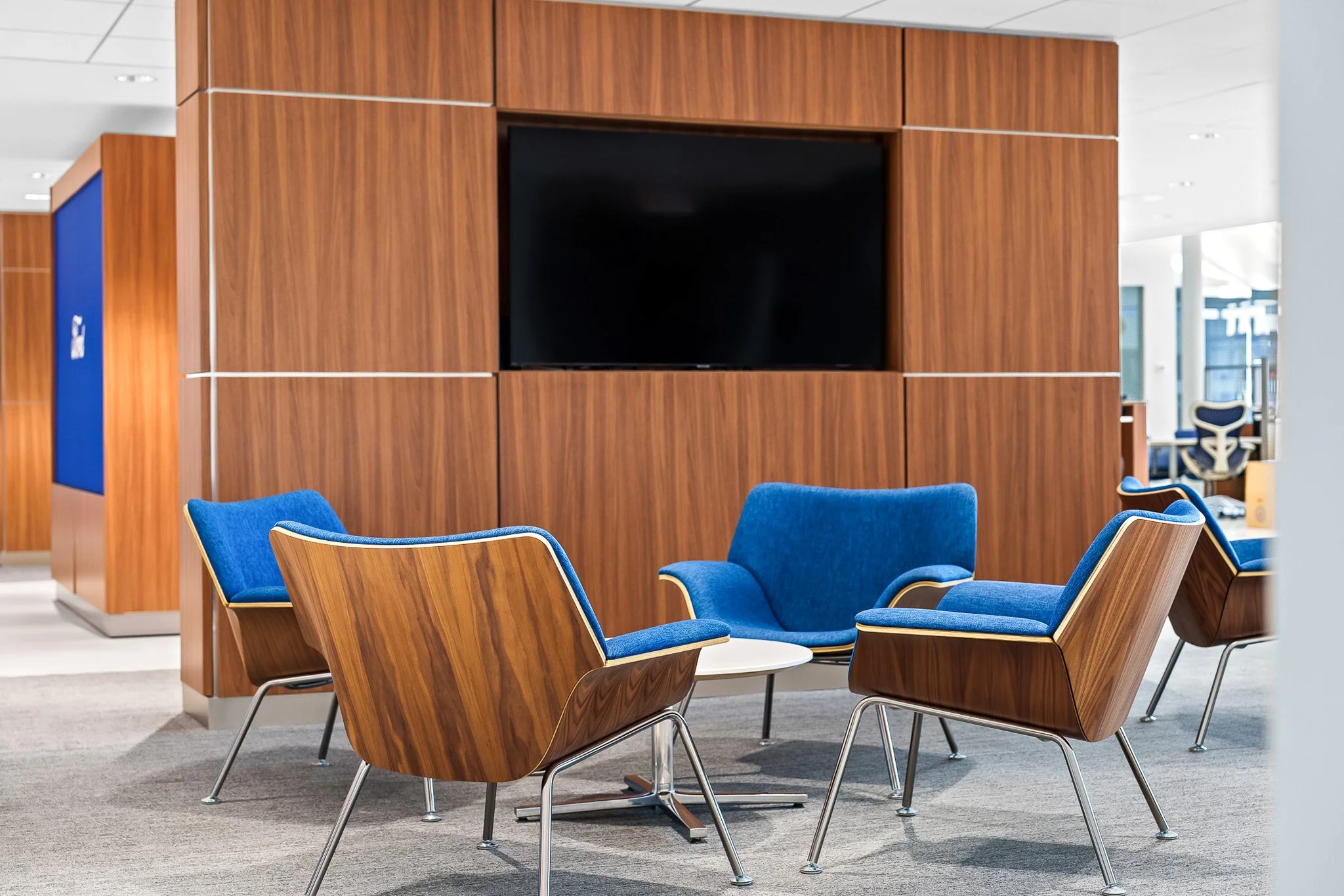 A modern office lounge area with blue cushioned chairs and a wooden partition with a large black screen mounted on it.