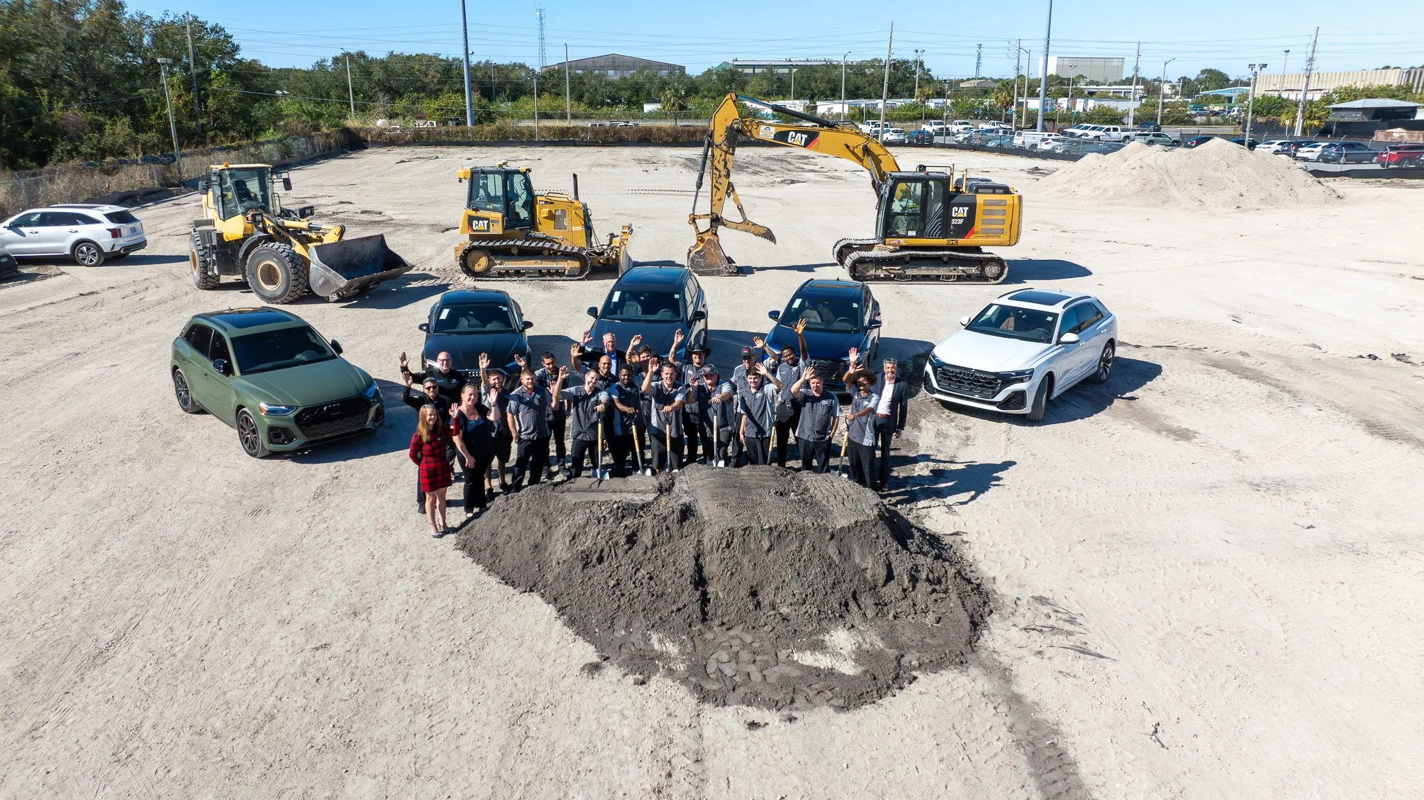 A large group of people, including men and women, standing together on a dirt construction site with shovels, posing for a photo. Behind them are several cars, construction equipment including excavators, and a partly cloudy sky.