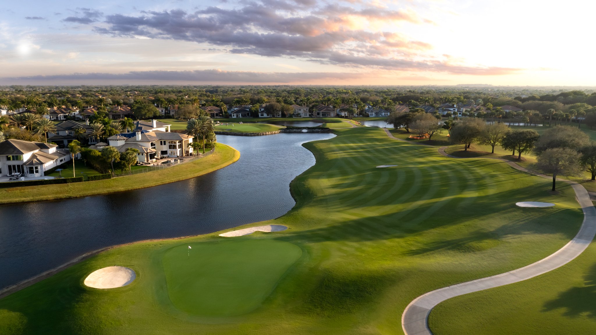 Aerial view of a golf course with neatly mowed green, sand bunkers, water hazards, walking path, trees, and residential houses in the background during sunset
