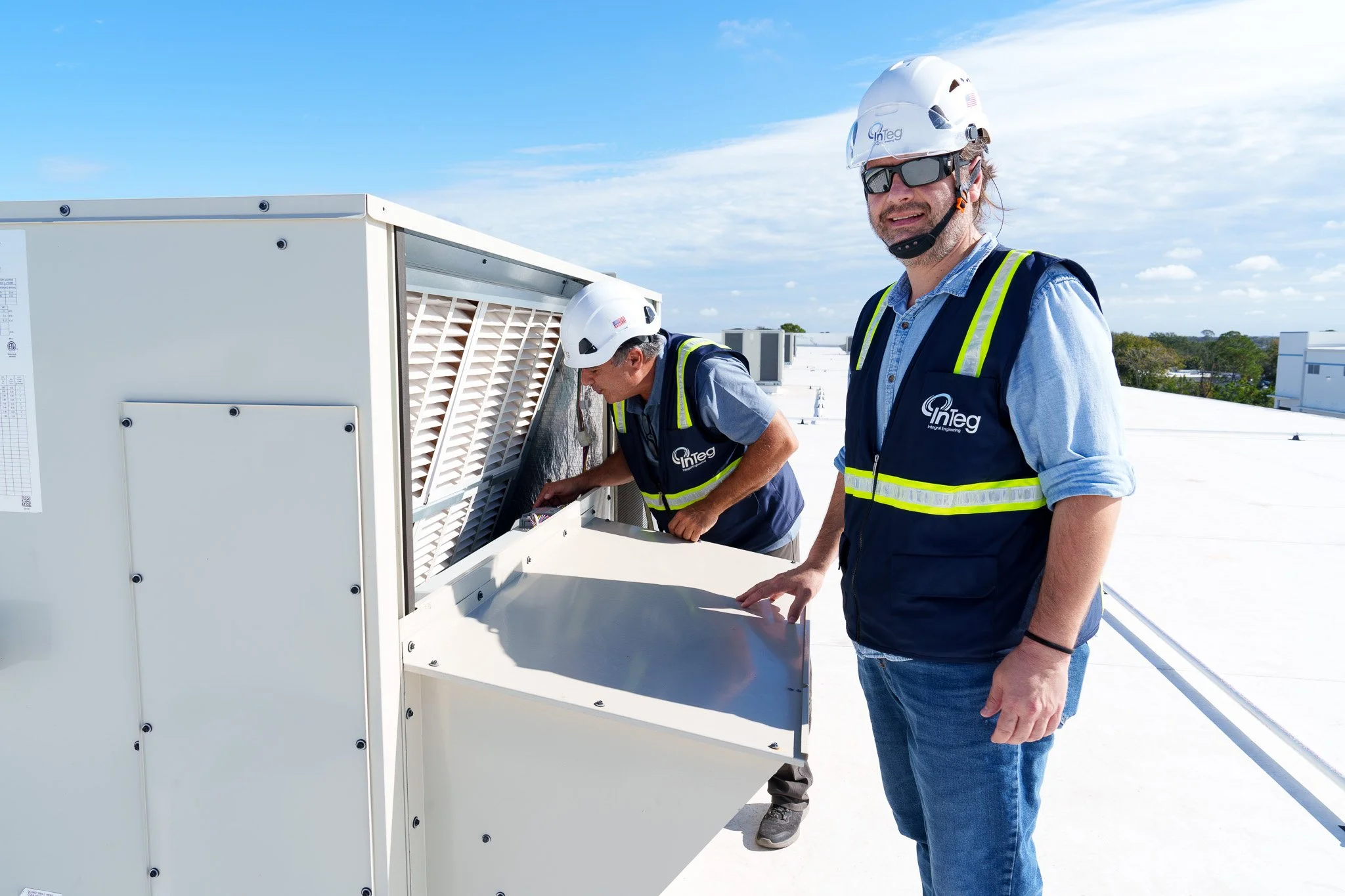 Two technicians wearing safety vests, helmets, and sunglasses working on an air conditioning unit on a rooftop under a blue sky.
