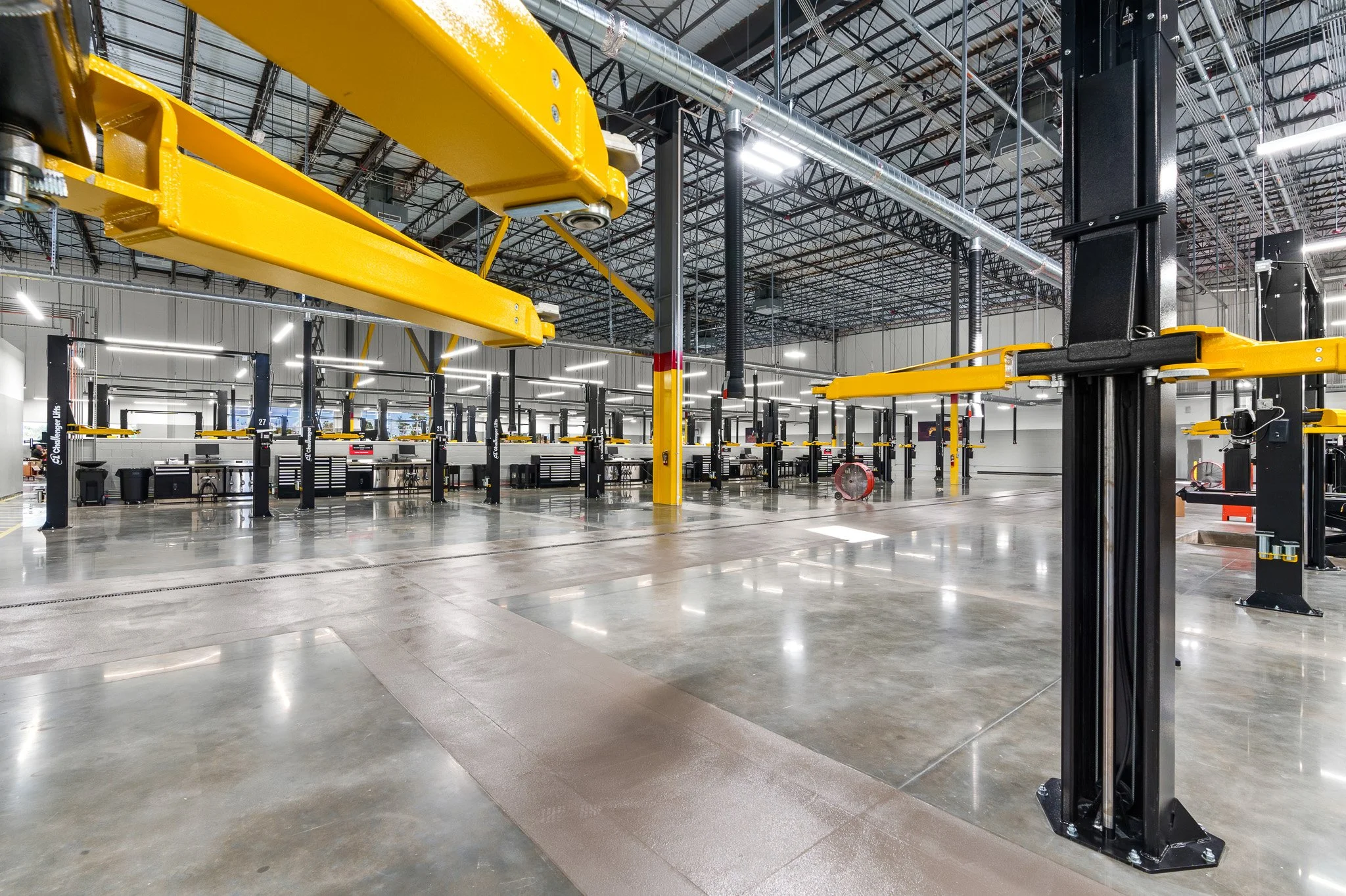 Inside of a vehicle repair shop with yellow overhead cranes, black lift supports, and a spacious, well-lit concrete floor.