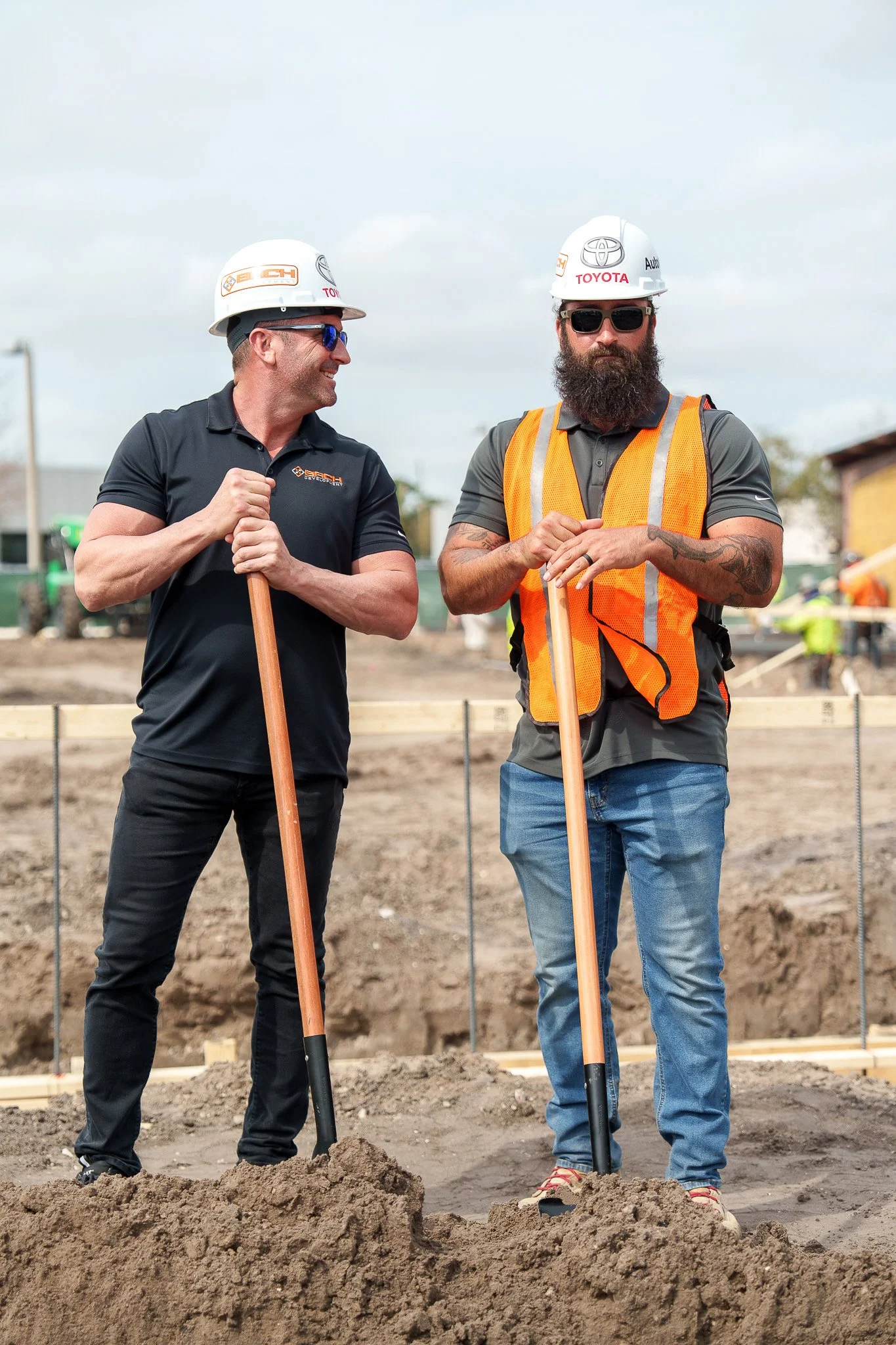 Two construction workers, one smiling and the other with a serious expression, standing on a construction site with shovels in hand. Both are wearing safety helmets and safety glasses. One is wearing a black polo shirt, and the other is wearing a gra