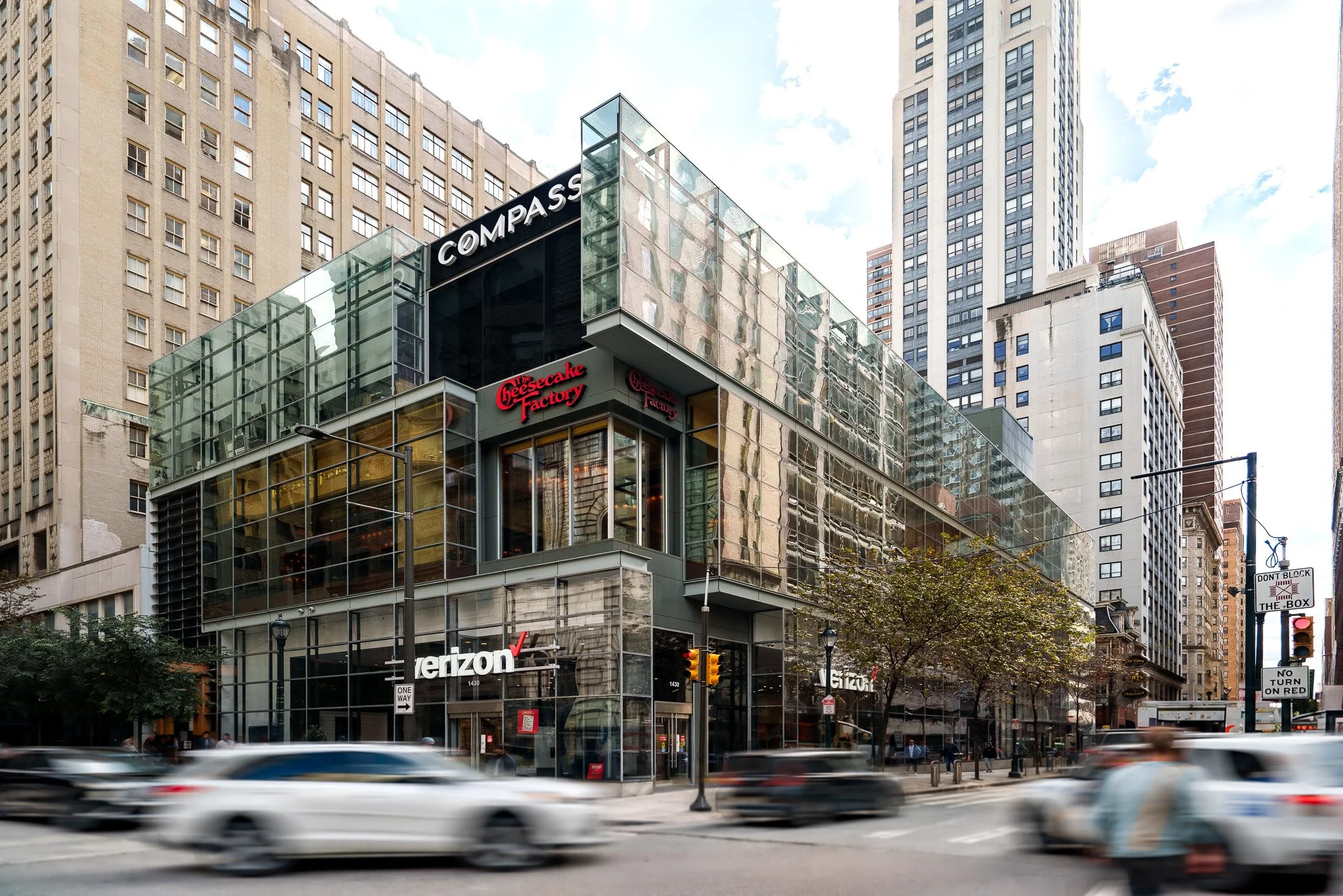 Urban street scene with a modern glass building housing Verizon and The Cheesecake Factory, surrounded by tall skyscrapers and via-moving cars in the foreground.