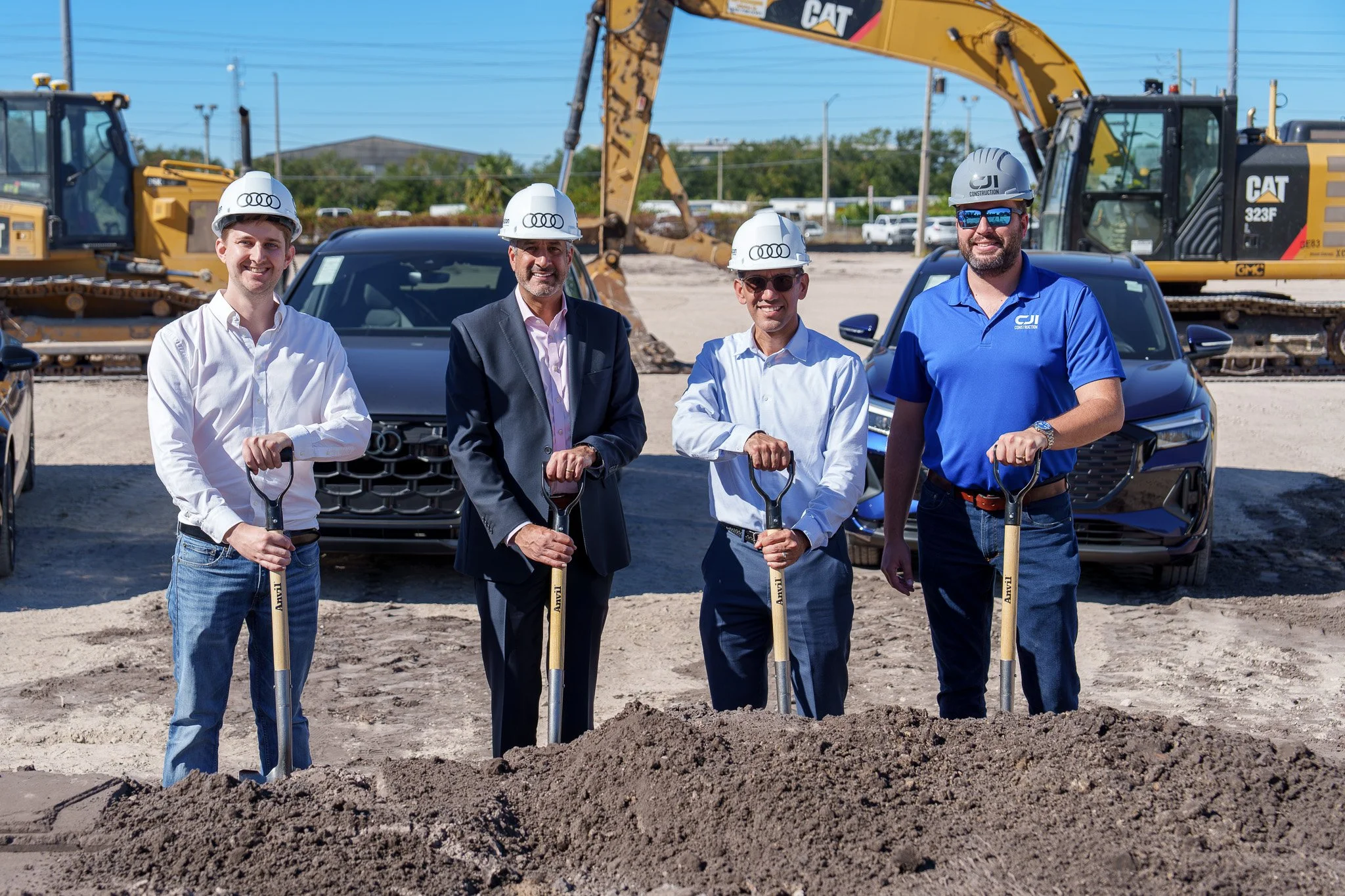 Four men in construction helmets and business attire holding shovels at a construction site with heavy machinery and parked cars in the background.