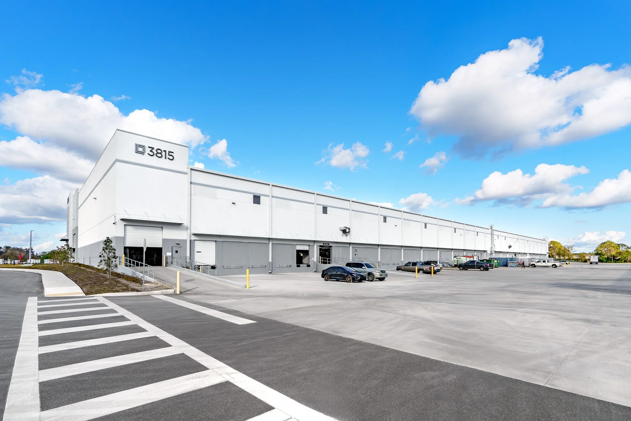 Large white warehouse building with the number 3815 on the side, parking lot with several cars, clear blue sky with scattered clouds.