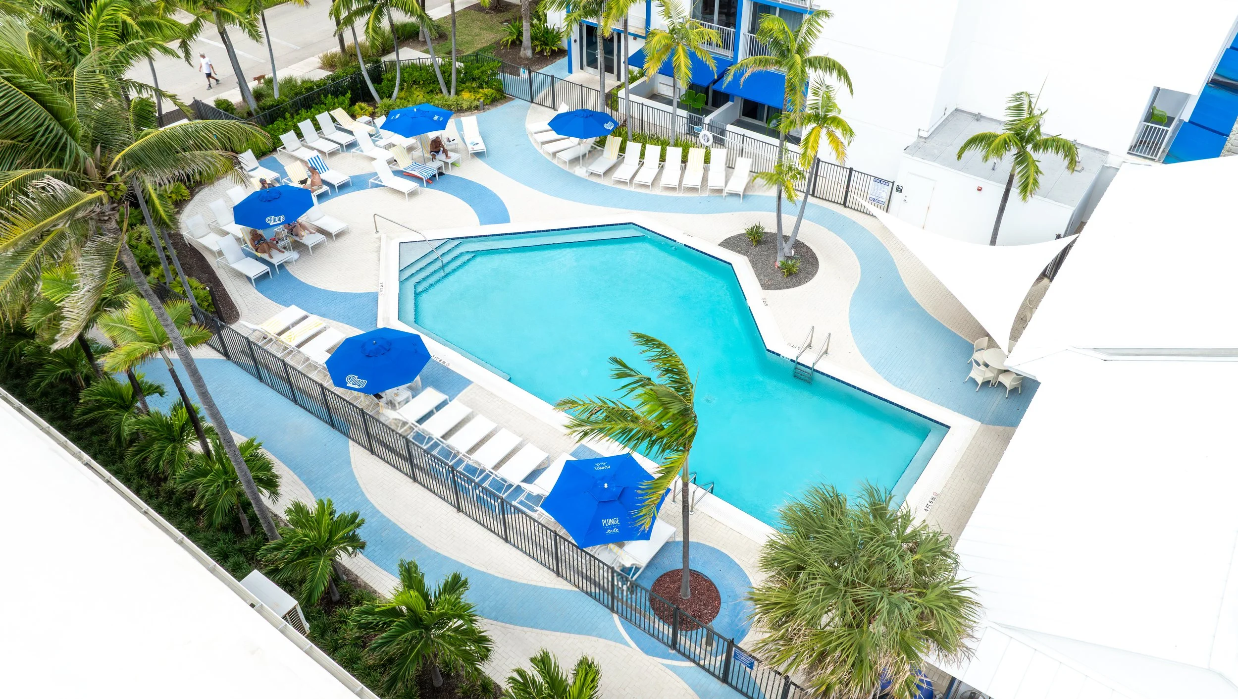 Elevated aerial photograph of a South Florida hotel pool deck with blue umbrellas and lounge chairs suitable for OTA listings and direct booking pages