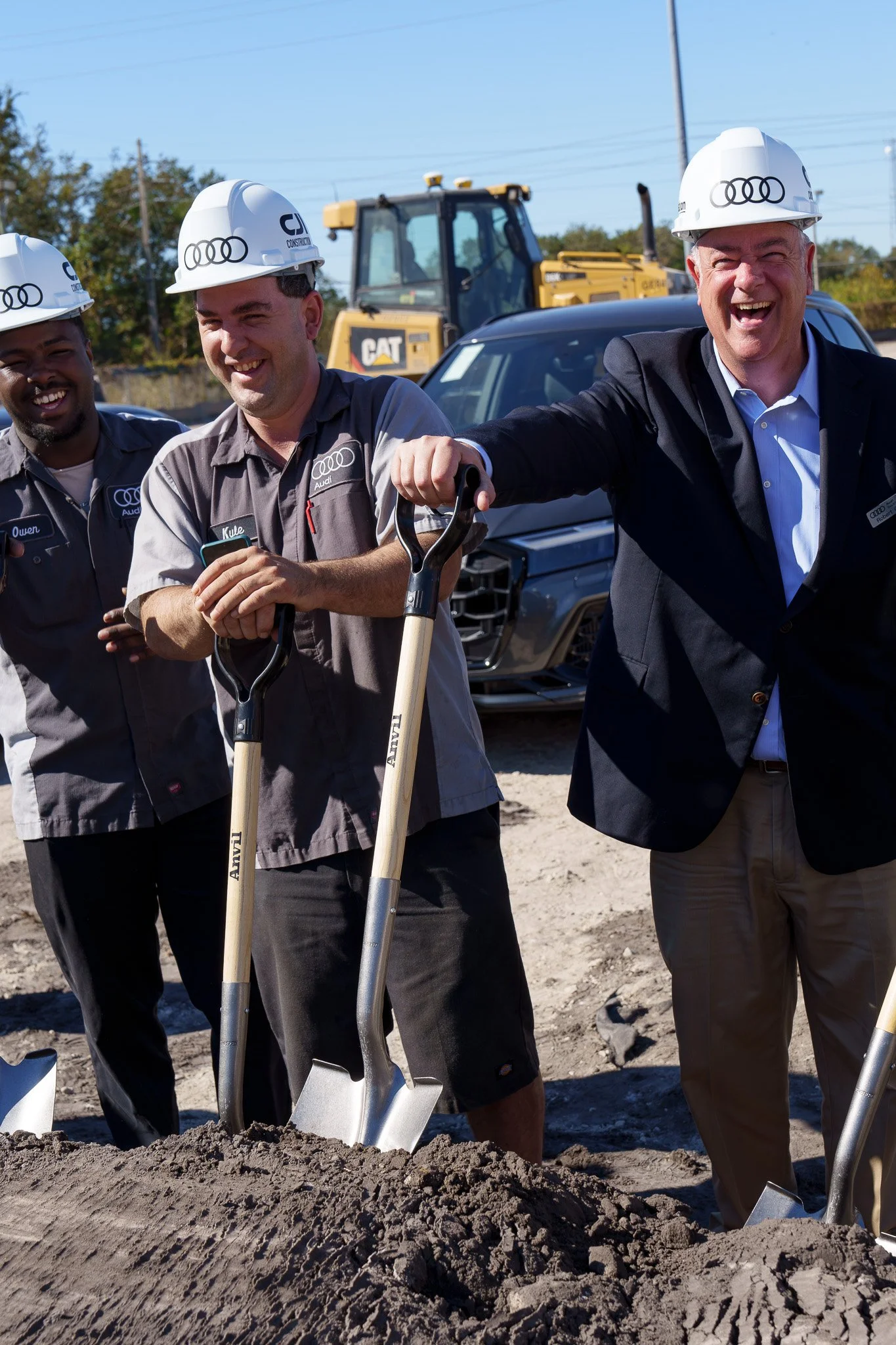 Three men wearing hard hats and work uniforms standing in a construction site, smiling, with a shovel in the ground and a yellow excavator in the background.