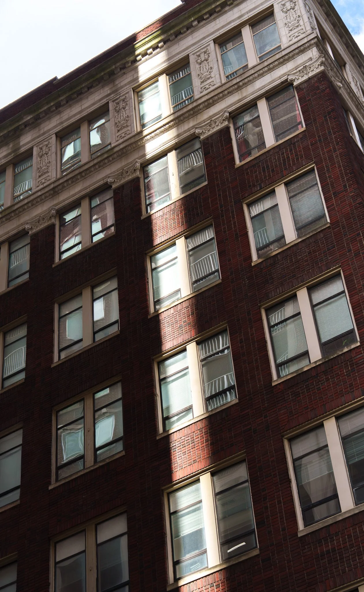 A tall brick apartment building with multiple windows, some of which have white railings or blinds. Sunlight partially illuminates the building, casting shadows on the facade.