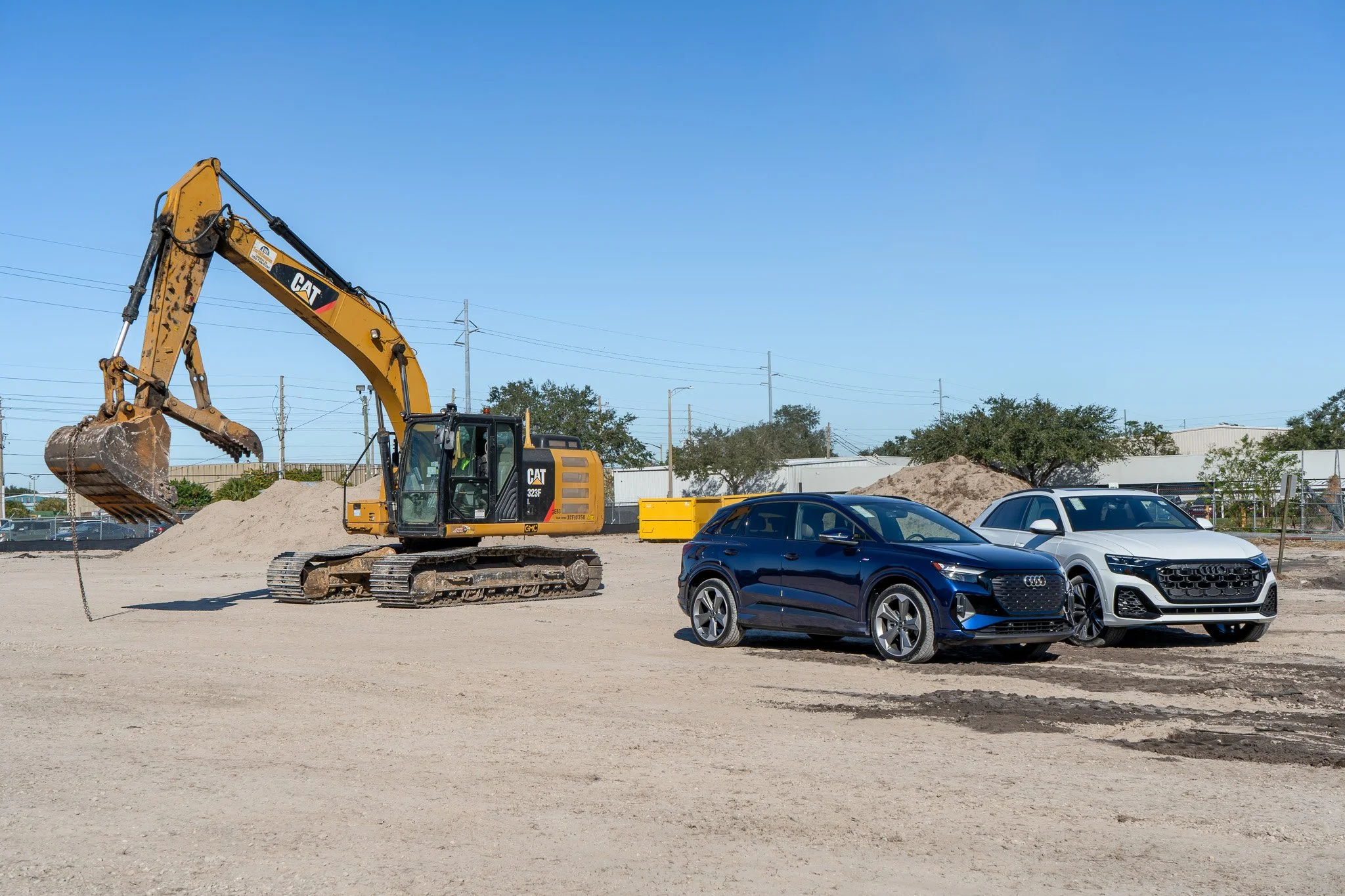 Construction site with a yellow Caterpillar excavator and several parked cars under a clear blue sky.
