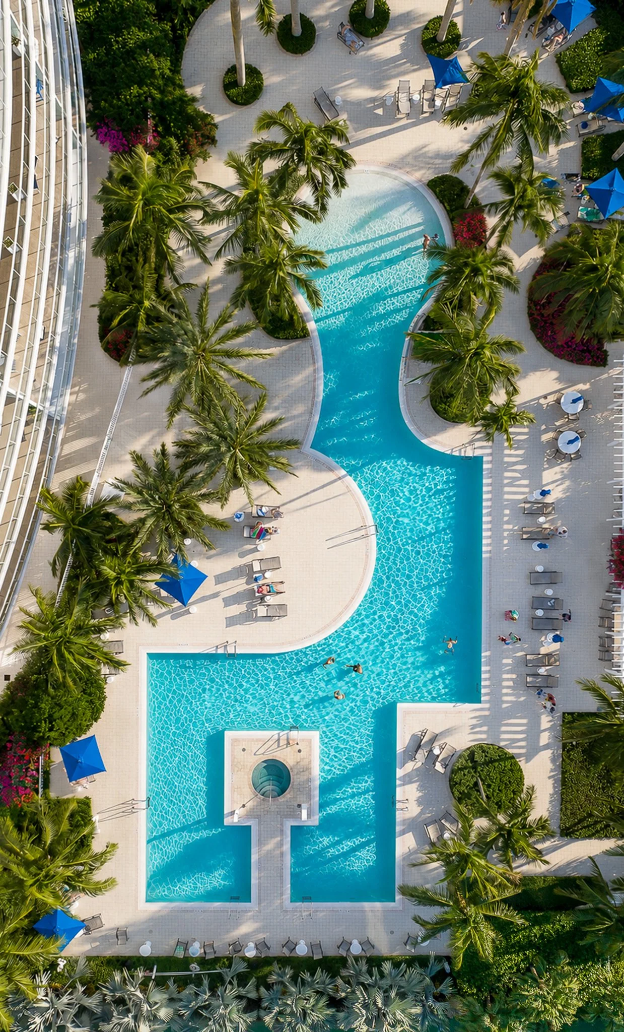 Aerial view of a hotel swimming pool surrounded by palm trees, lounge chairs, and umbrellas in a tropical setting.