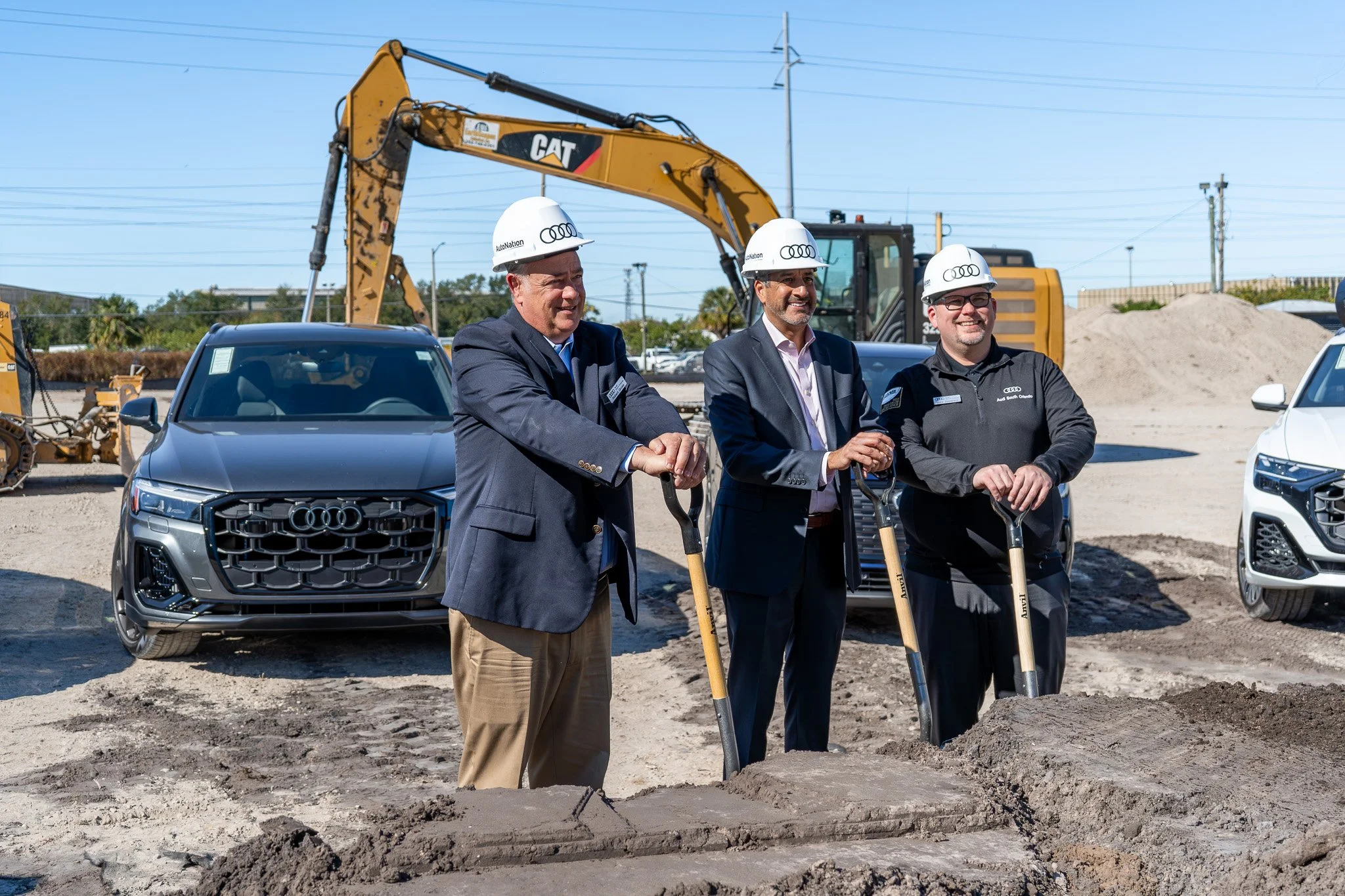 Three men in suits and helmets stand on a construction site, smiling and posing with shovels, with construction equipment and cars in the background.