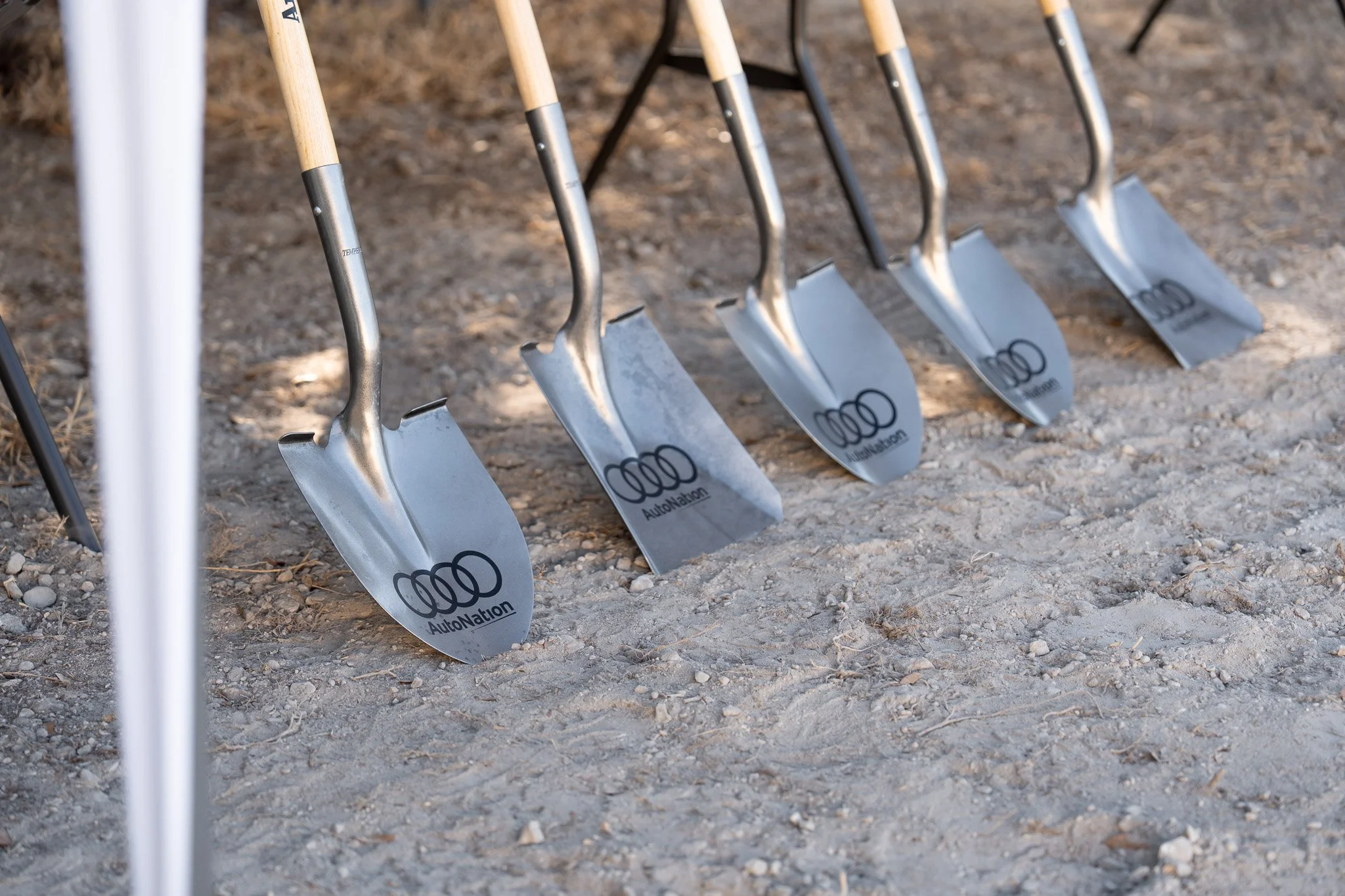 A row of metal shovels with wooden handles, labeled with the Audi logo and 'AutoNation,' standing upright on a gravel surface.