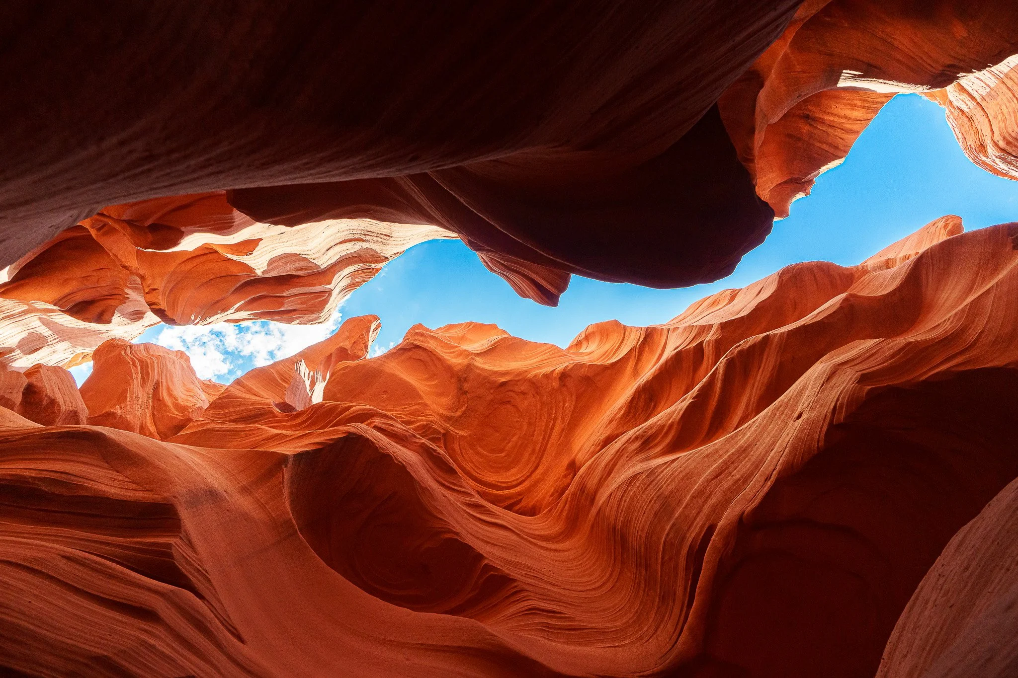 View inside a narrow slot canyon with smooth, wavy sandstone walls in shades of orange and red, and a patch of blue sky visible at the top.