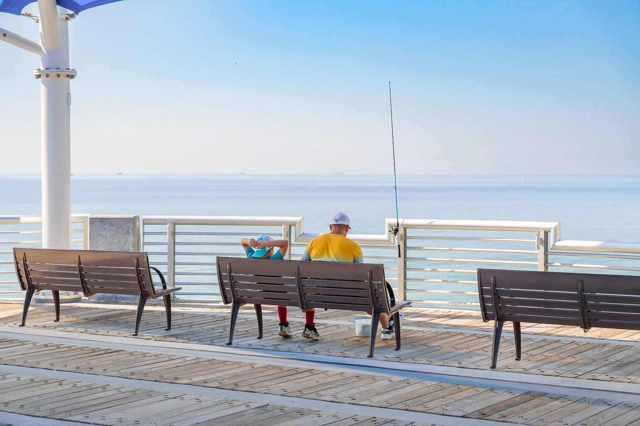 Two people sitting on benches on a pier, next to the ocean, with a fishing pole nearby. The person on the left is wearing a blue shirt and cap, while the person on the right is wearing a yellow shirt, white cap, and red socks.