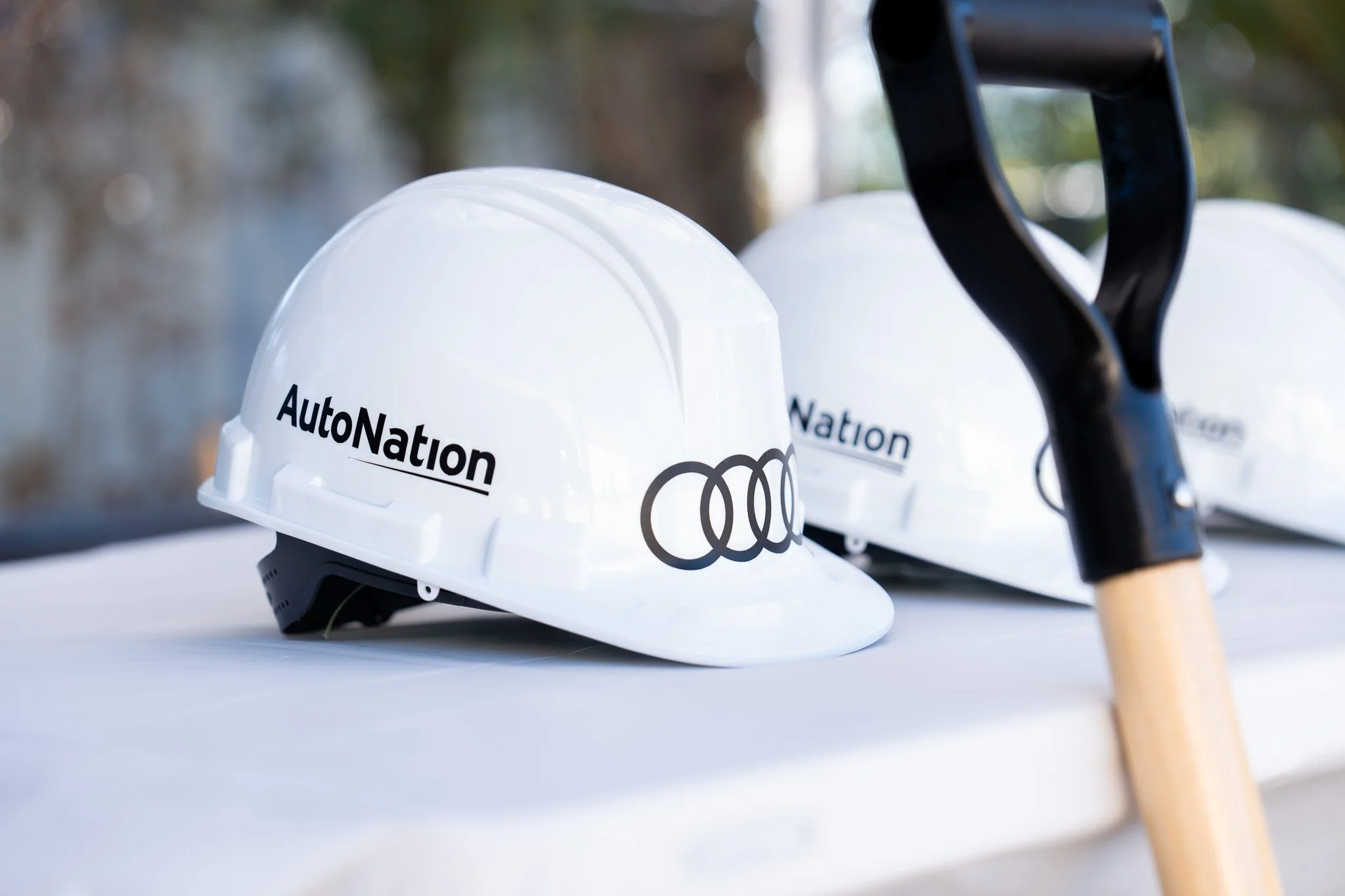 Two white helmets with AutoNation logo and Audi rings sitting on a white surface, with a kicking tee and a wooden tee in front of them, and a blurred outdoor background.