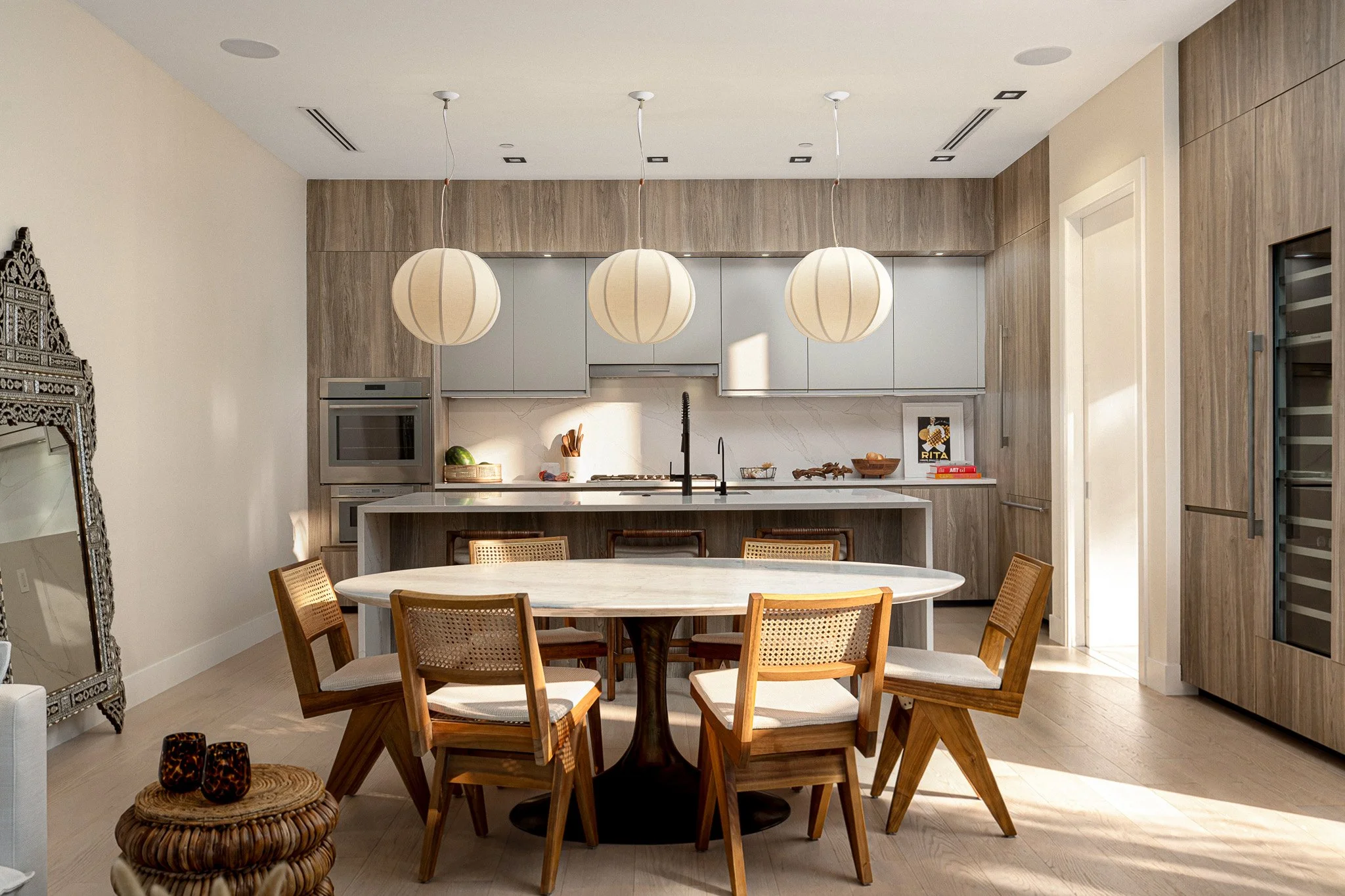 Modern kitchen with a white round dining table surrounded by wooden chairs, three large pendant lights hanging above, wooden cabinetry, a built-in oven, and decorative items on the counter.