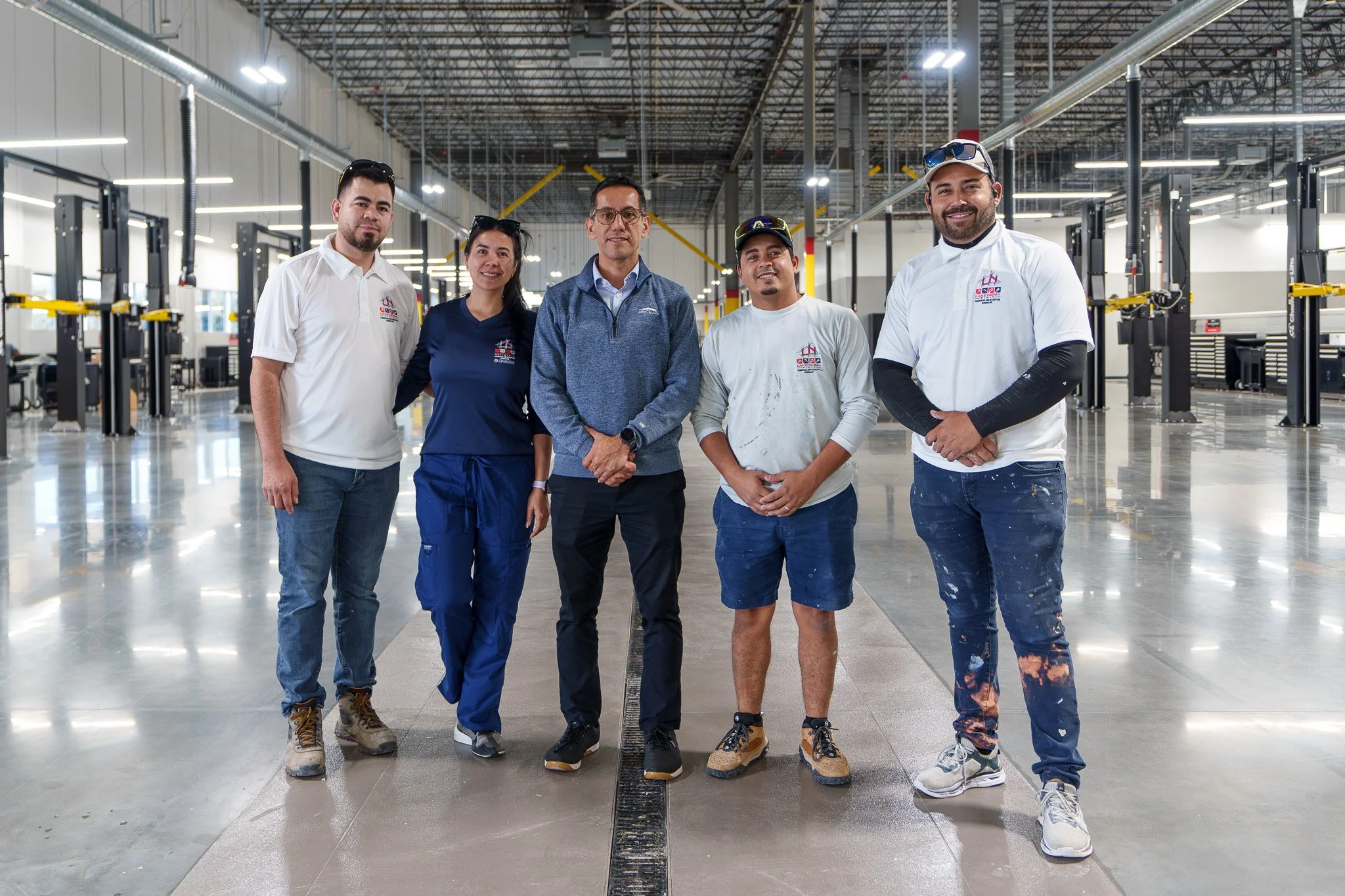 Group of five diverse workers standing inside a large industrial or manufacturing facility, smiling at the camera.