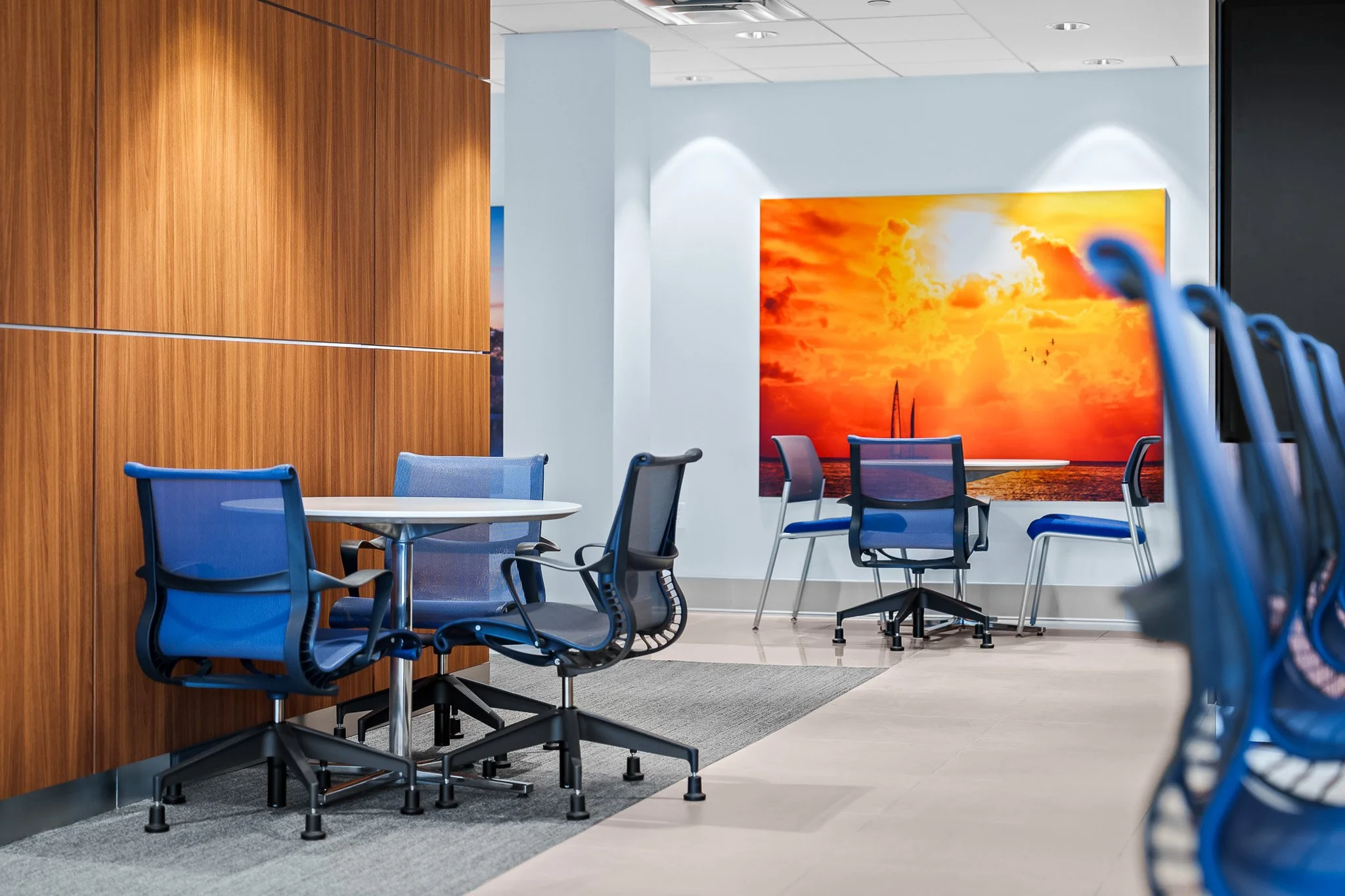 Conference room with blue office chairs, a large orange sunset artwork, wooden wall panels, and a gray carpet.