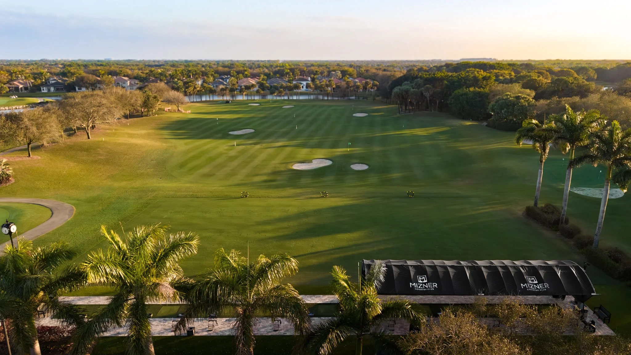 A scenic view of a golf course with well-maintained green fairways, sand bunkers, and trees, with residential houses in the distance, under a partly cloudy sky during sunset.