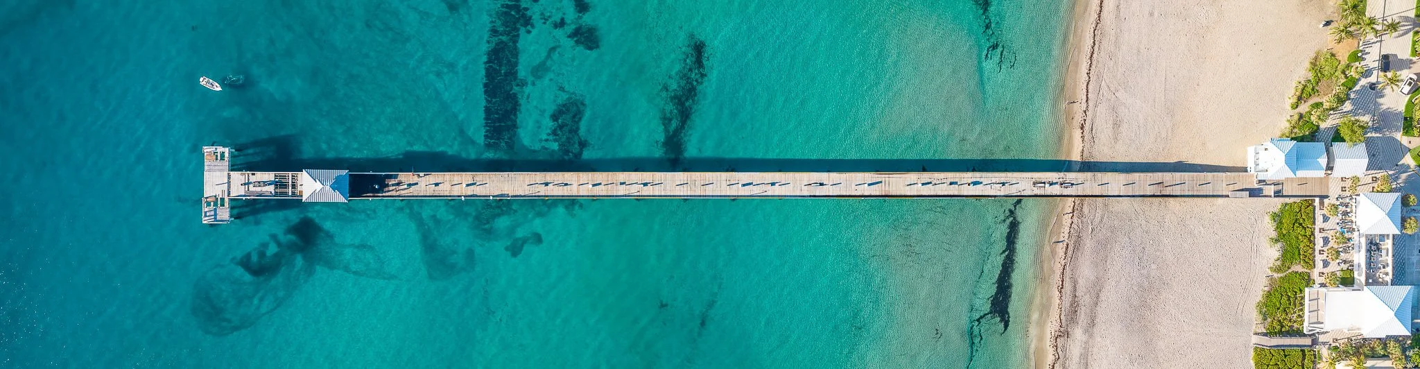 An aerial view of a long pier extending over turquoise ocean water towards a sandy beach with houses and palm trees.