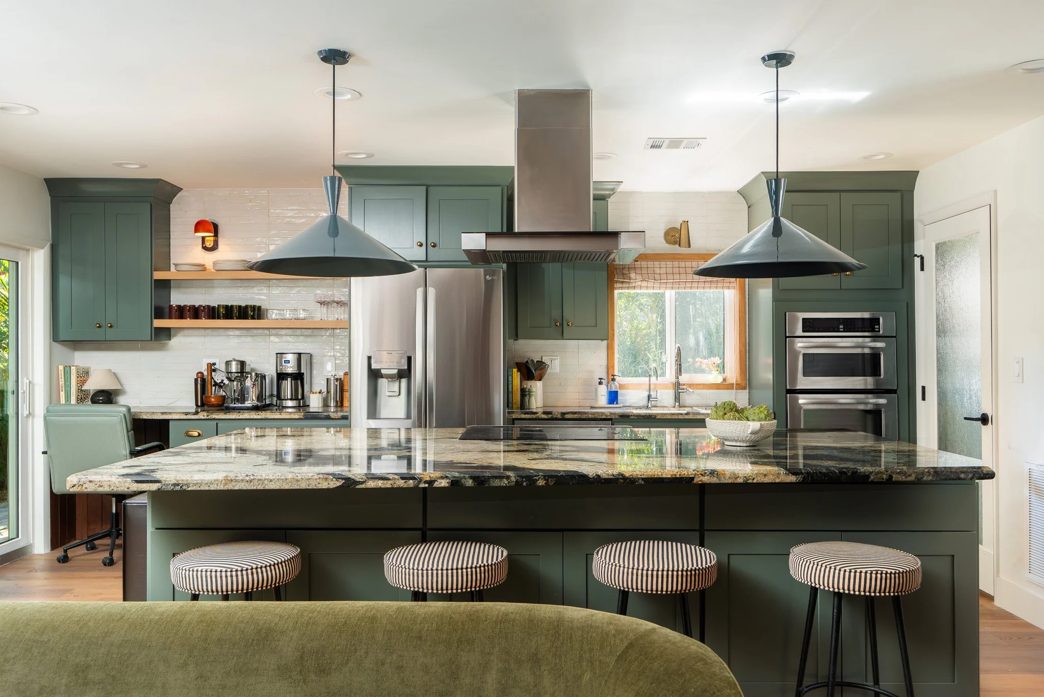Modern kitchen with green cabinetry, granite island, pendant lights, stainless steel appliances, open shelves, and a window with a red checkered curtain.