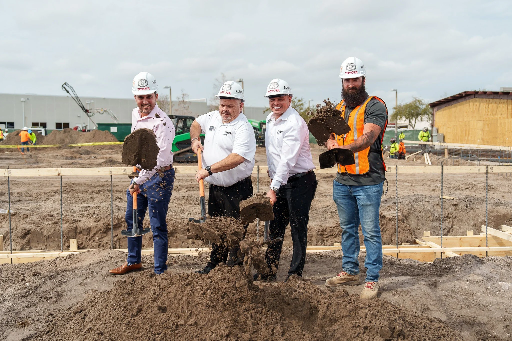 Four men, wearing hard hats and work clothes, are at a construction site pouring dirt into a trench. They are smiling and holding shovels. there are construction workers in the background.