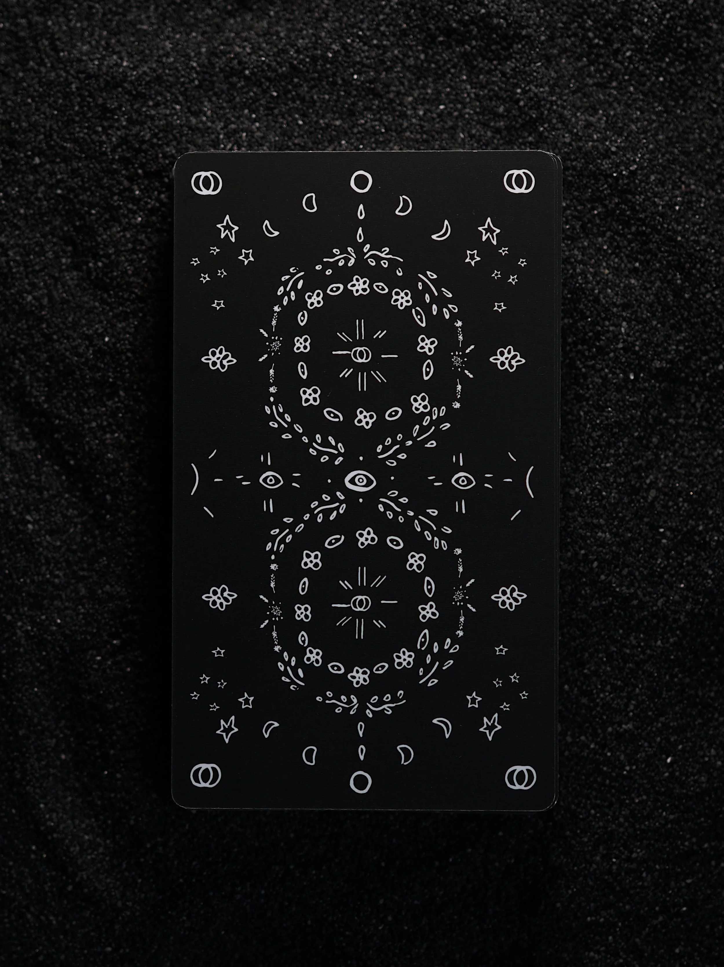 A black tarot card with white abstract mystical symbols and patterns, including circles, stars, moons, and eyes, laid on a black textured surface.