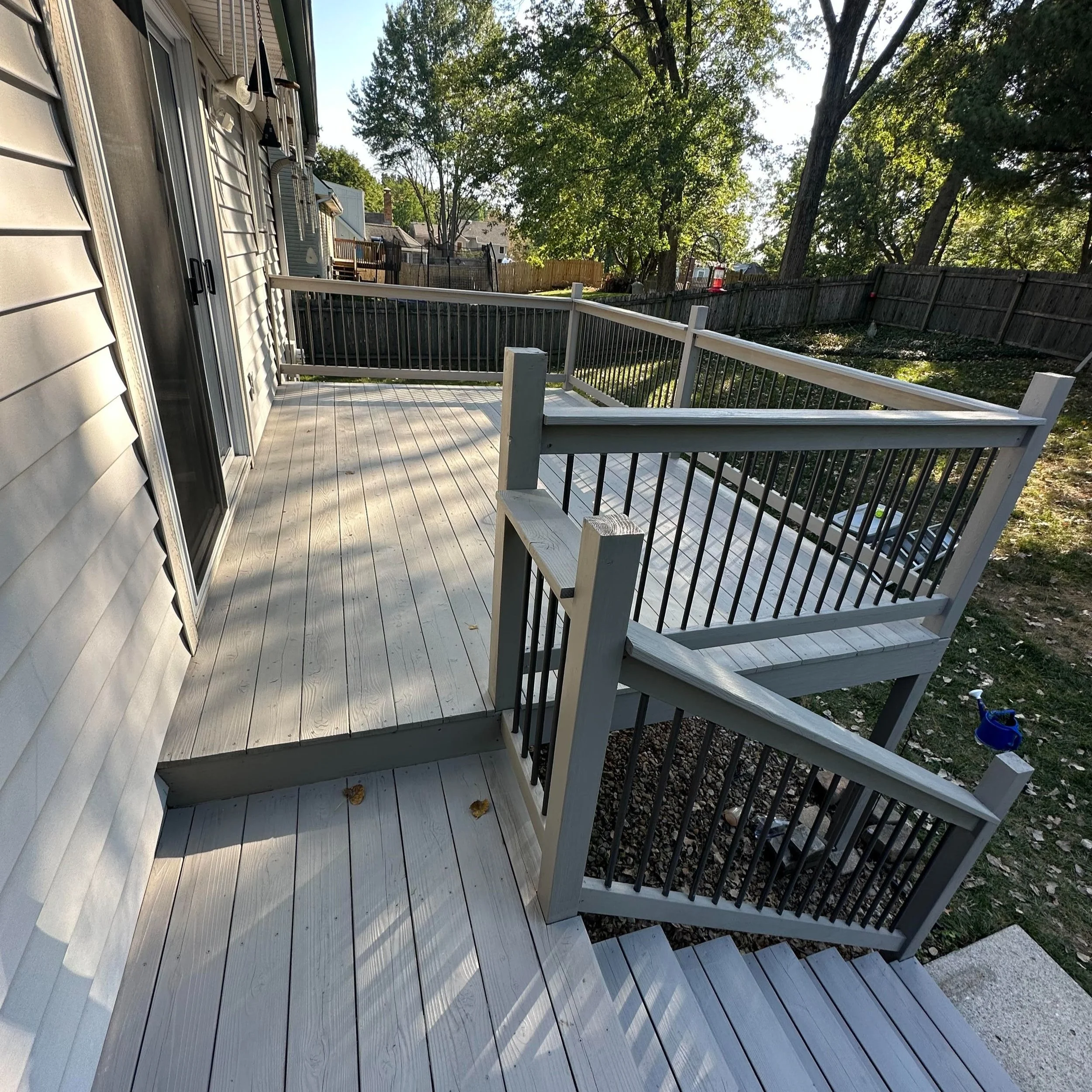 Deck with wooden floorboards, surrounded by a white and black metal railing, connected to stairs leading to a grassy backyard with trees and a wooden fence.