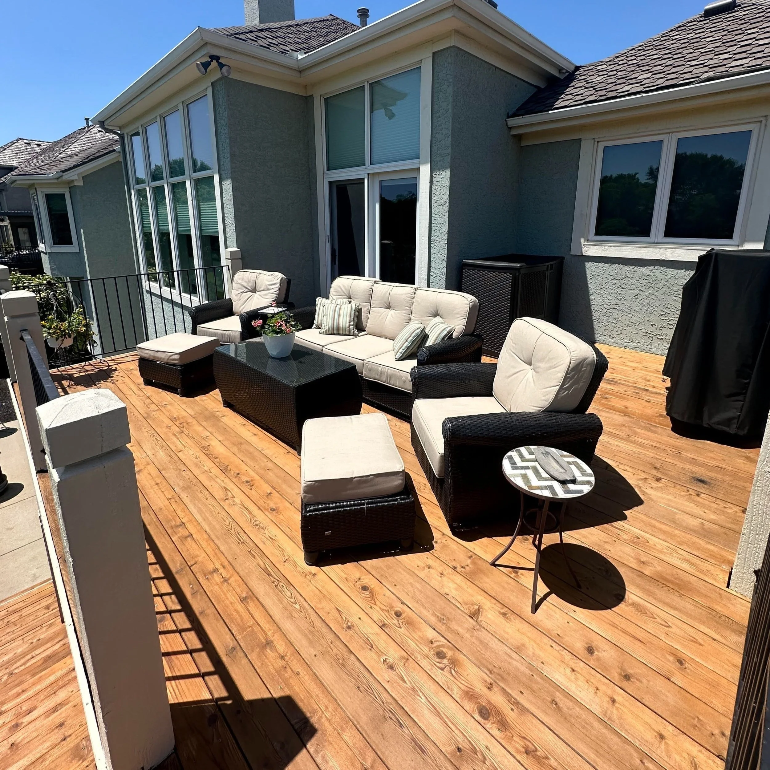 Outdoor wooden deck with beige cushioned wicker furniture including a sofa, two armchairs, an ottoman, and a small round side table with a zigzag patterned top, situated against a light green house with multiple windows and a sliding glass door, under bright sunshine.