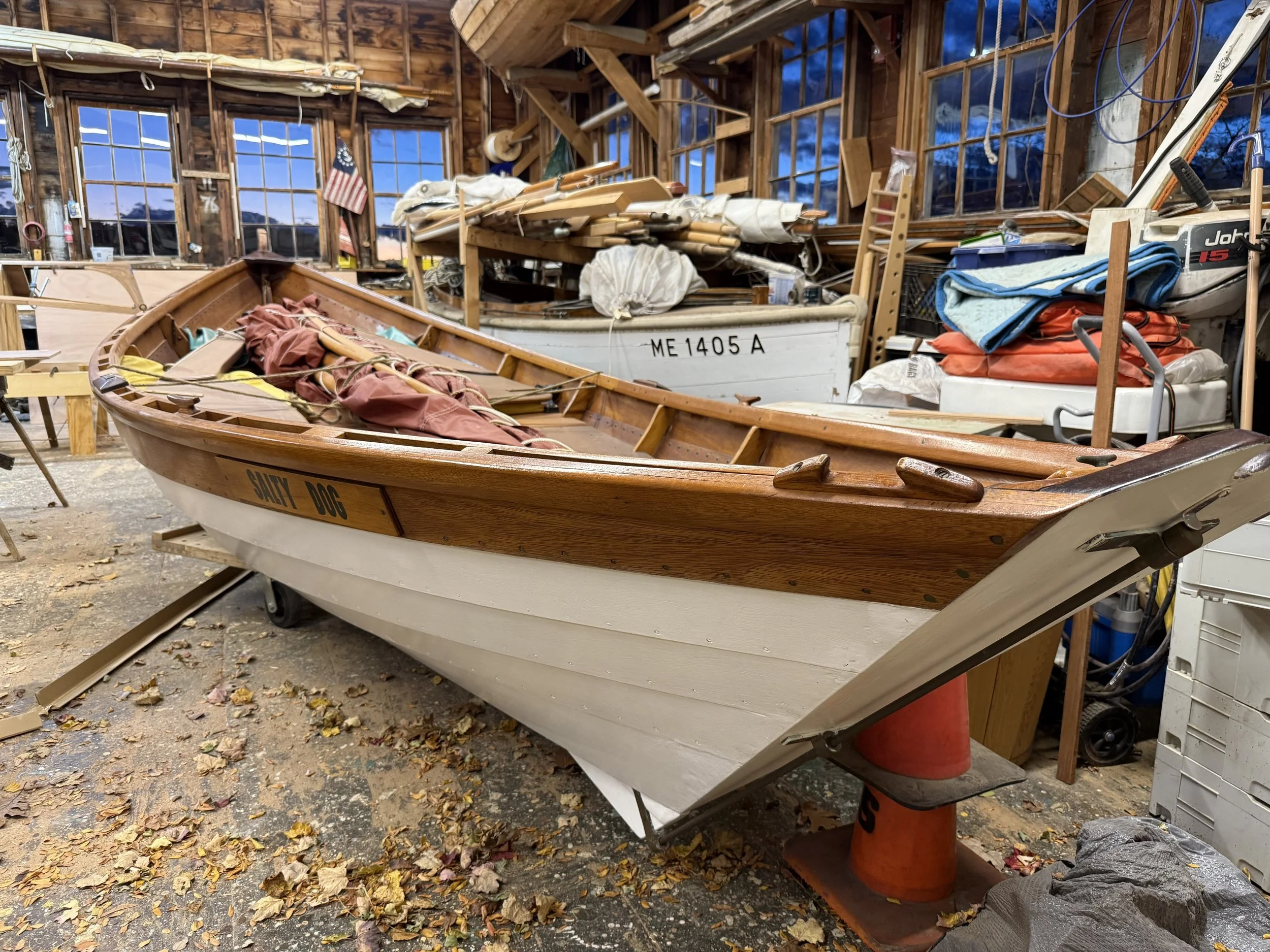 A 19' white wooden lapstrake dory from the stern looking forward with a varnished shearstrake and rails, filled with sailing gear, rudders, and oars. The shop floor in the foreground has leaves and sawdust.