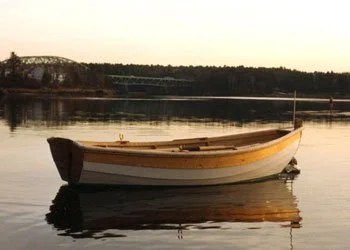 A lowell tender, white with varnished sheer strake, in late evening light sits in the calm water of the Merrimack River with a bridge in the background.