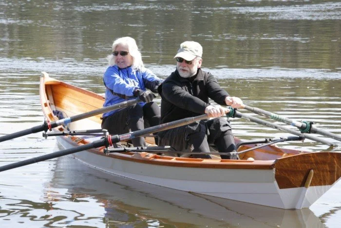 Two people in sliding rowing seats in an atlantic skiff with white topsides, a buff sheer strake, and varnished transom and gunwales.