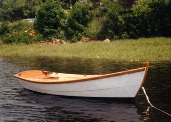 A sleek Deer Island Skiff with its elegant sheerline and white topsides floats at a mooring with a grassy bank and woods in the background.