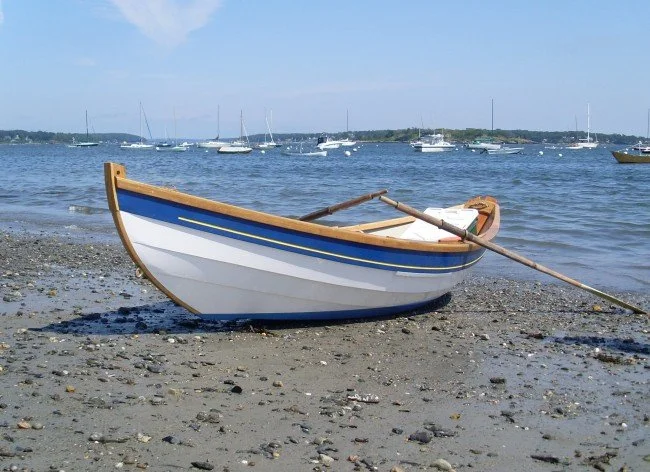 A surf dory with a blue sheerstrake, varnished gunwales, and white topsides sits on a sandy beach with boats in a mooring in the background on a sunny day.