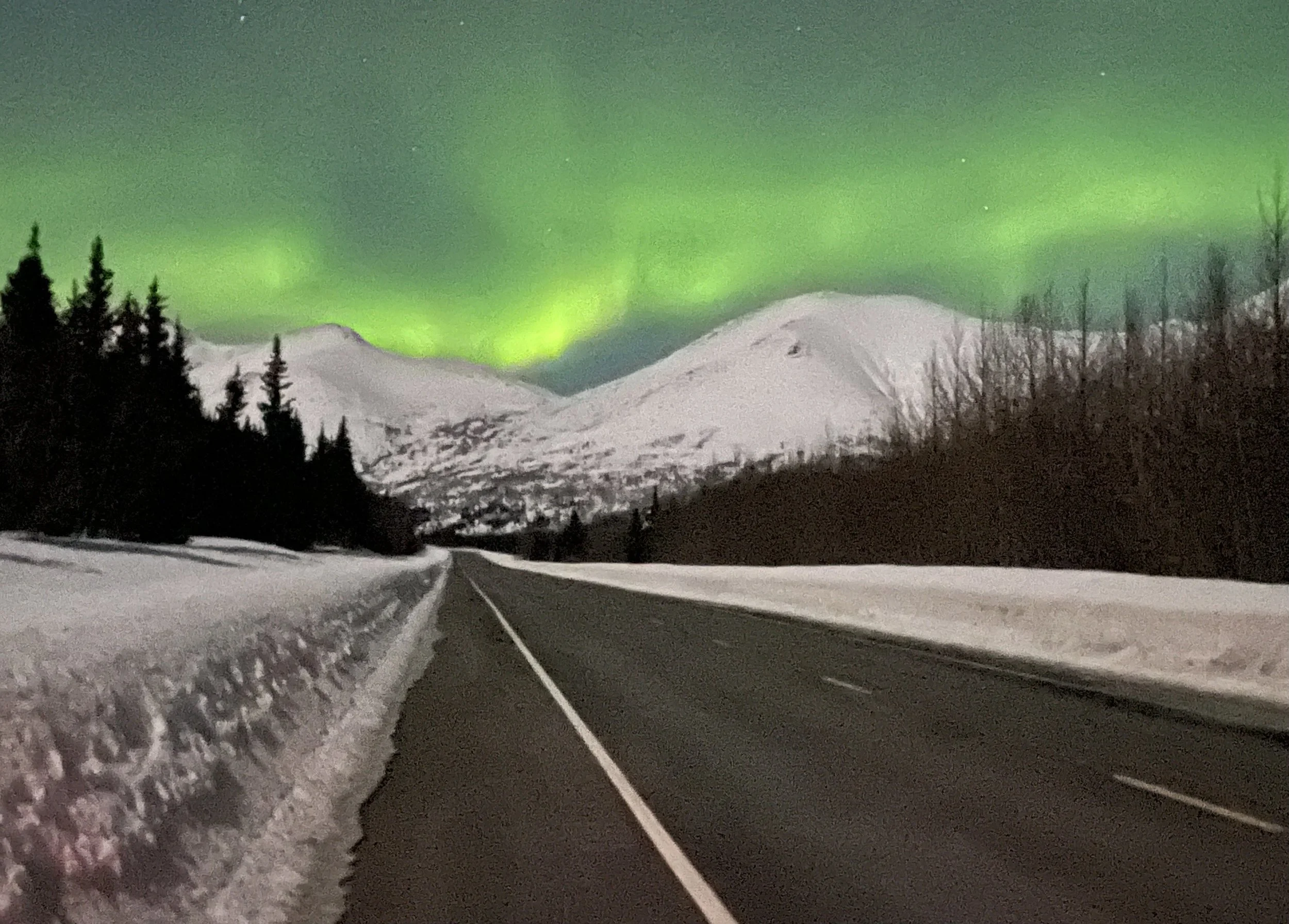 The northern lights or Aurora Borealis is seen above a straight road and the snow covered mountains towering over the road