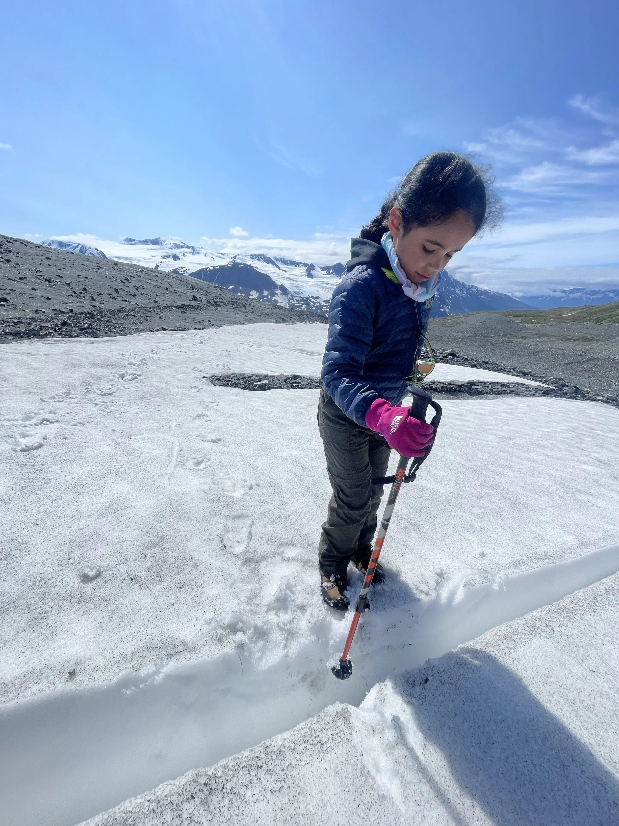 A young girl dressed in winter clothes and holding a trekking pole look into a small crevasse 