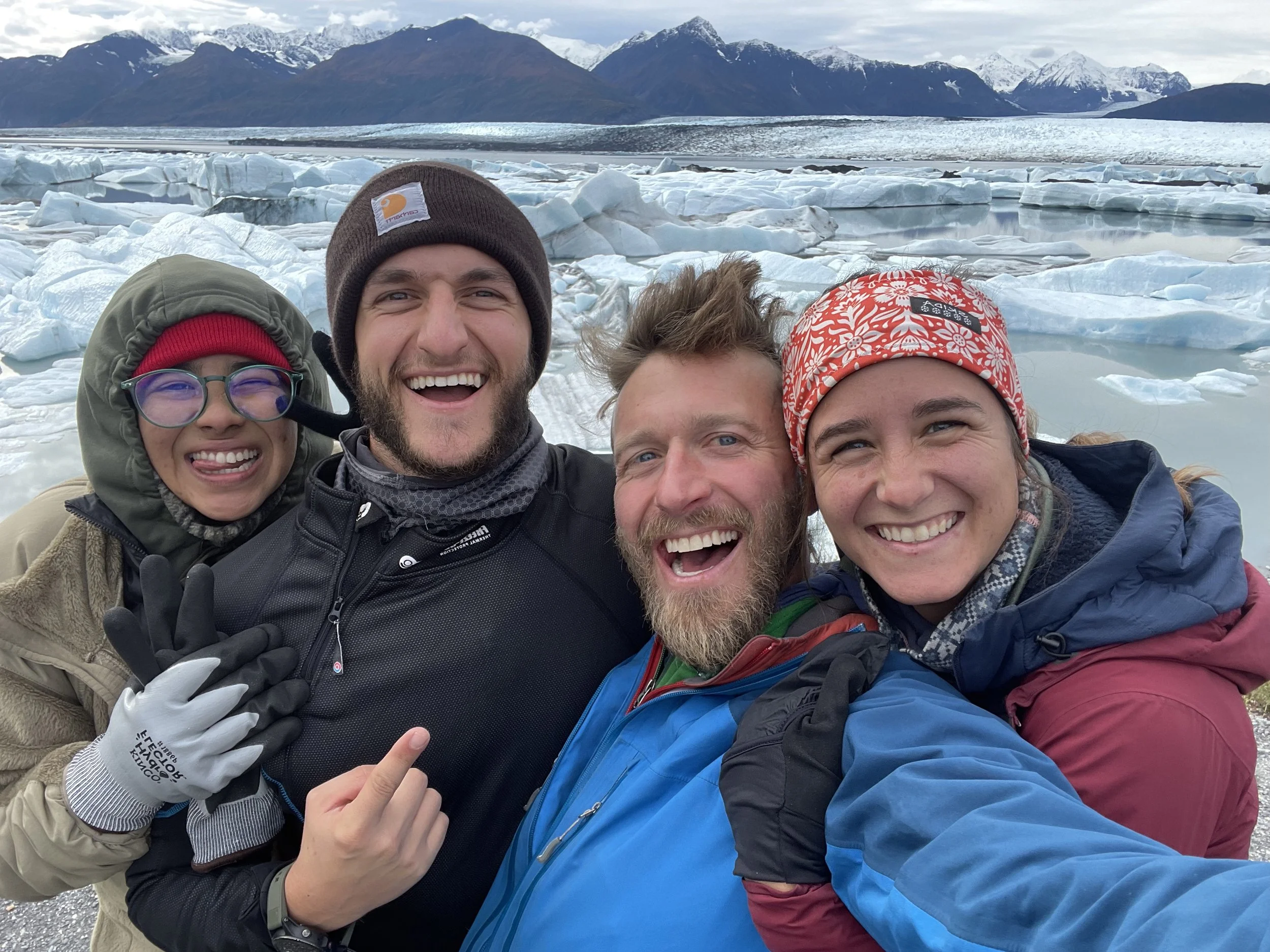 4 people laugh as they stand close together wearing winter clothing and pose in front of the Knik Glacier