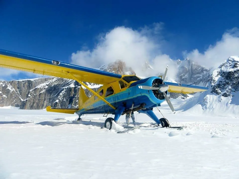 A yellow and blue bush plane sits on a glacier on a sunny day