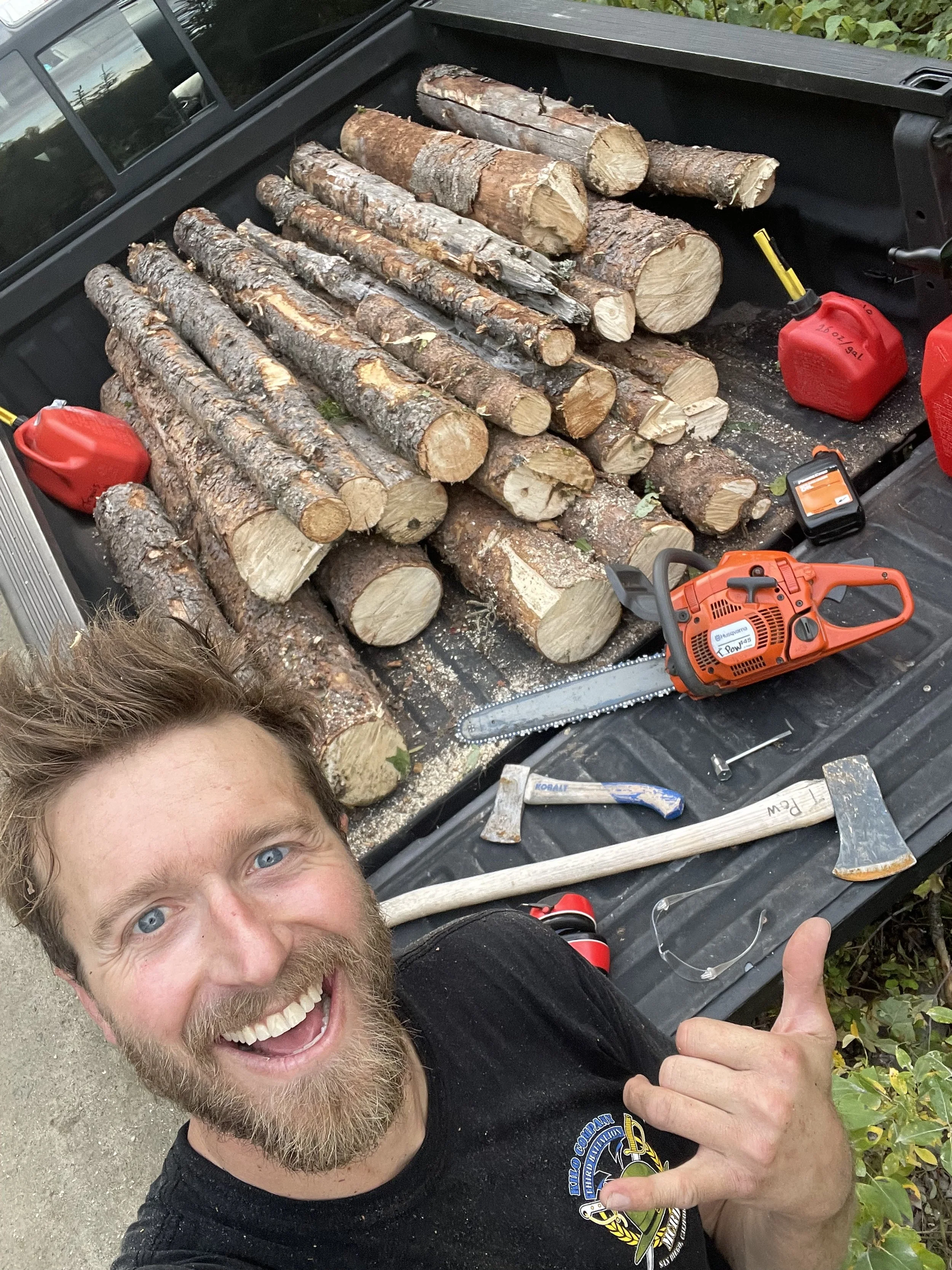 A man smiles in front of a truck loaded with cut wood, fuel cans, a chainsaw and wood splitting axes. 