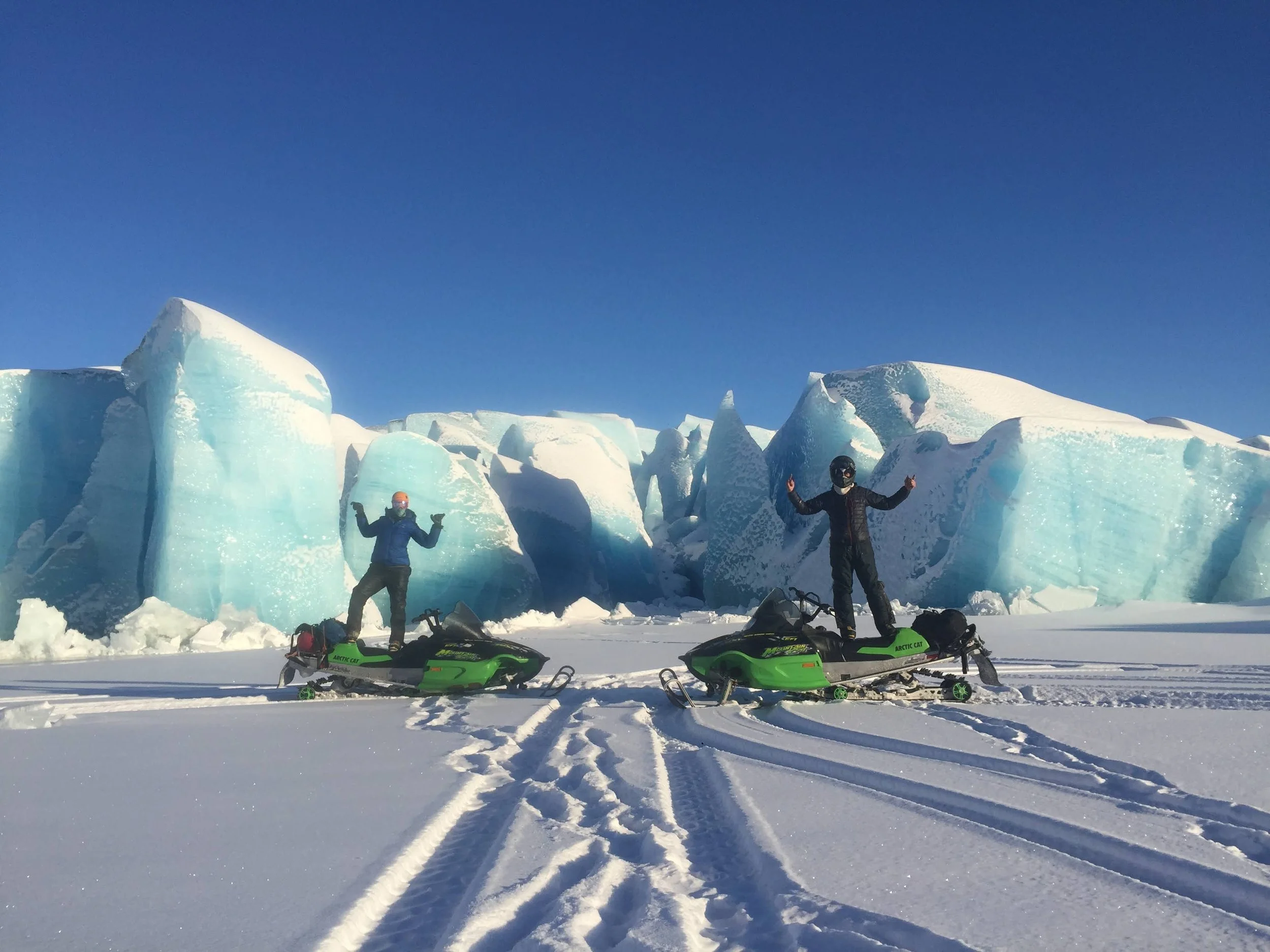 Two people stand on their snowmobiles and pose for a picture with their hands in the air. The snowmobiles are parked in front of large blue ice chunks from a glacier. 