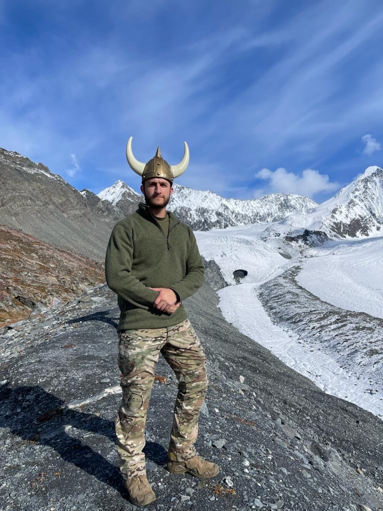 A man stands on the moraine of the Matanuska glacier wearing camo pants and a viking helmet