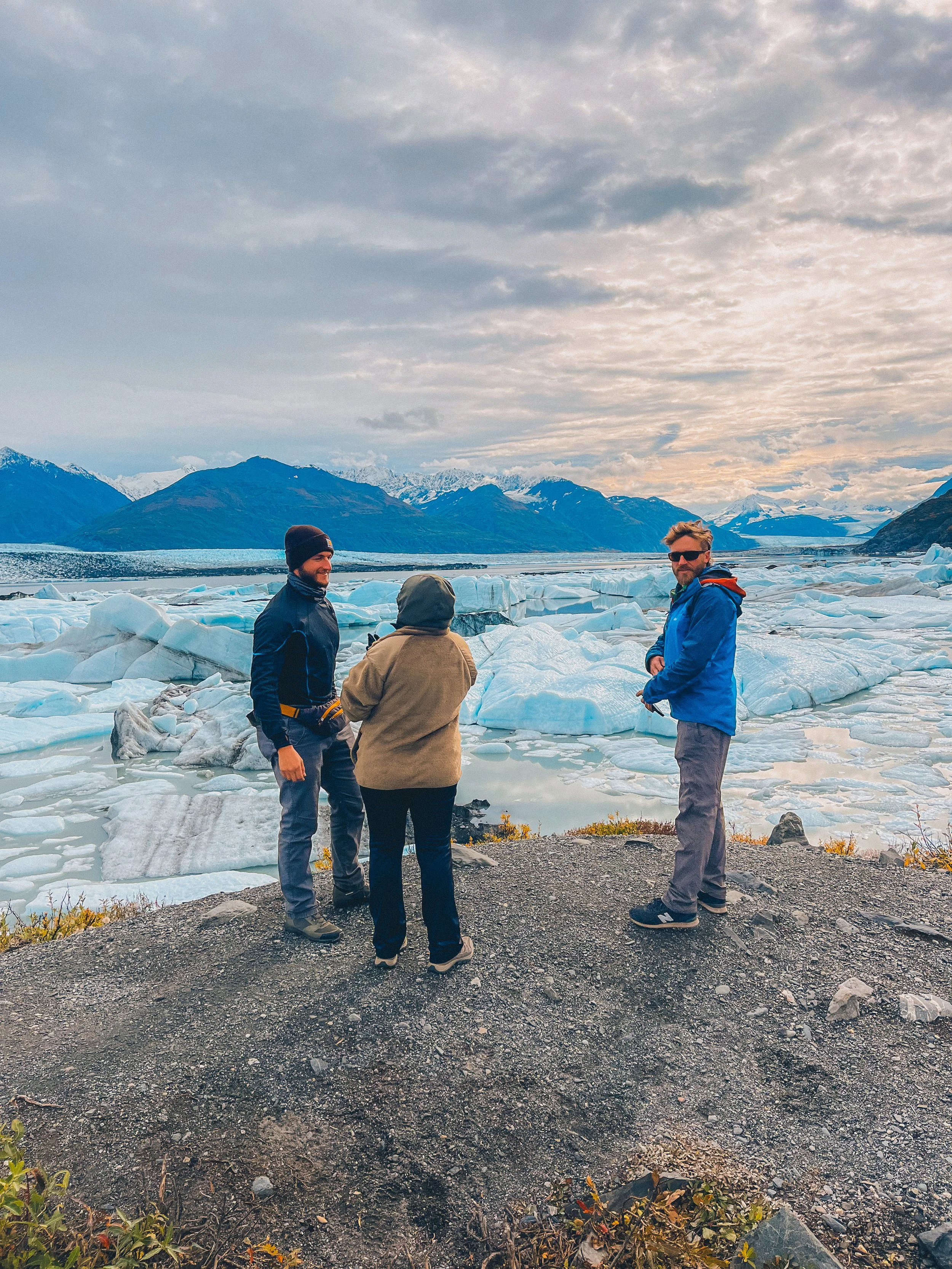 Three people stand together laughing in front of the glacier blue of icebergs that are floating in the water of the Knik river 