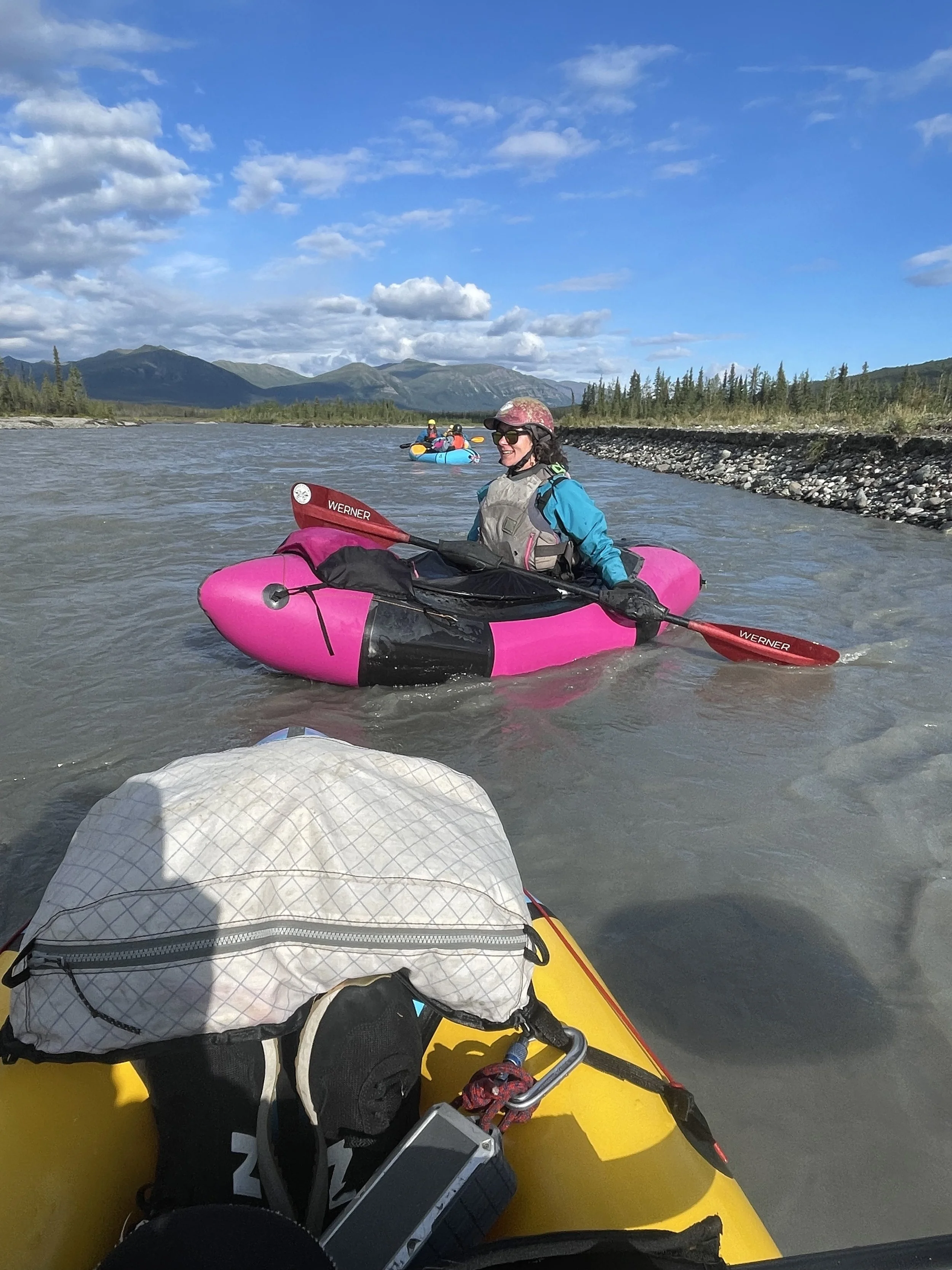 The person taking the photo sits in a yellow packraft while looking at a woman sitting in a pink packraft and paddling downstream on a sunny day.