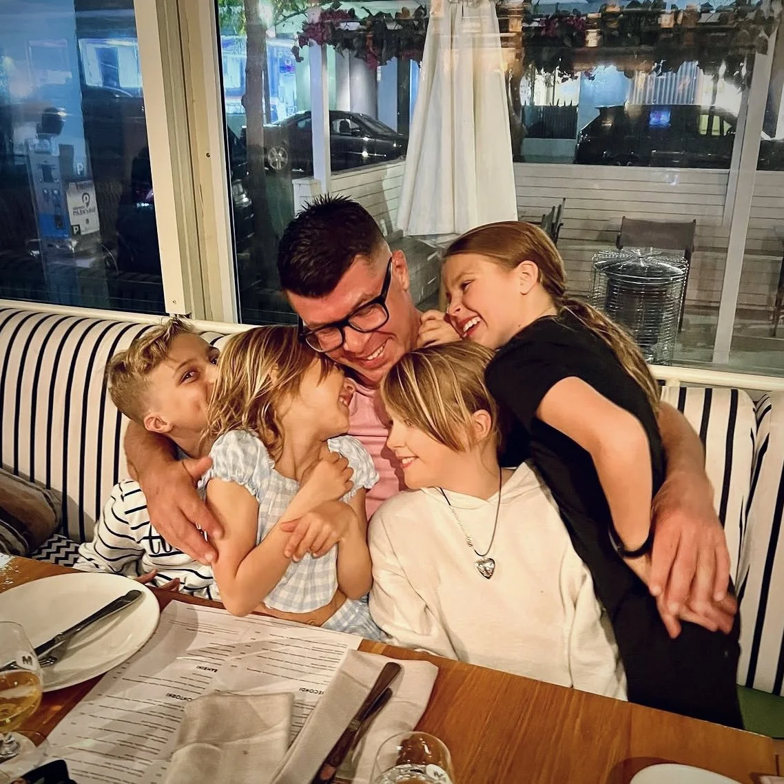A man surrounded by four children, all smiling and laughing, hugging each other joyfully in a restaurant setting.