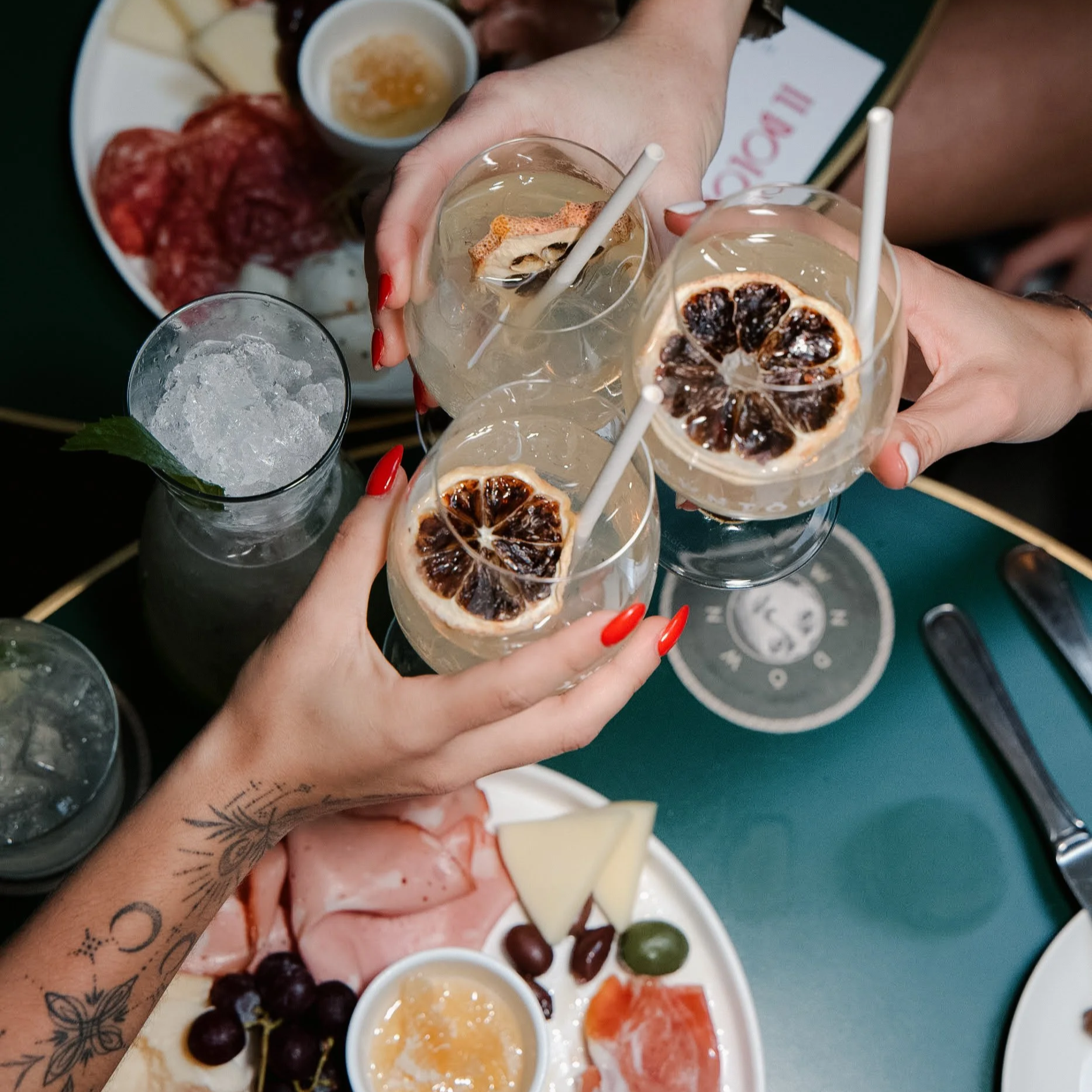 Multiple hands holding glasses of some beverage topped with slices of dried citrus fruit, with a tray of cheese, meats, grapes, and small bowls of sauces or dips in the background.
