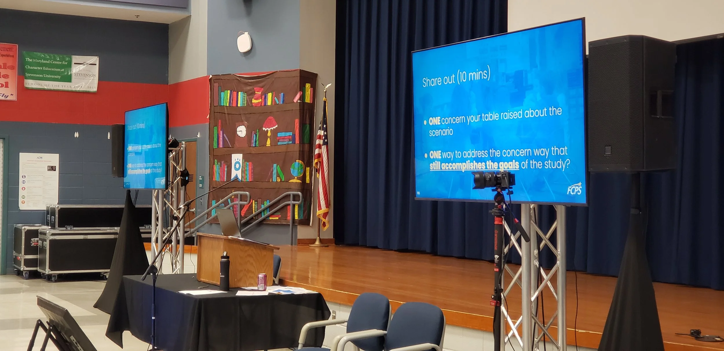 Presentation stage with large screen displaying instructions for a discussion, a camera on a tripod, blue curtains, and a decorated bulletin board with books and a clock in a school auditorium.