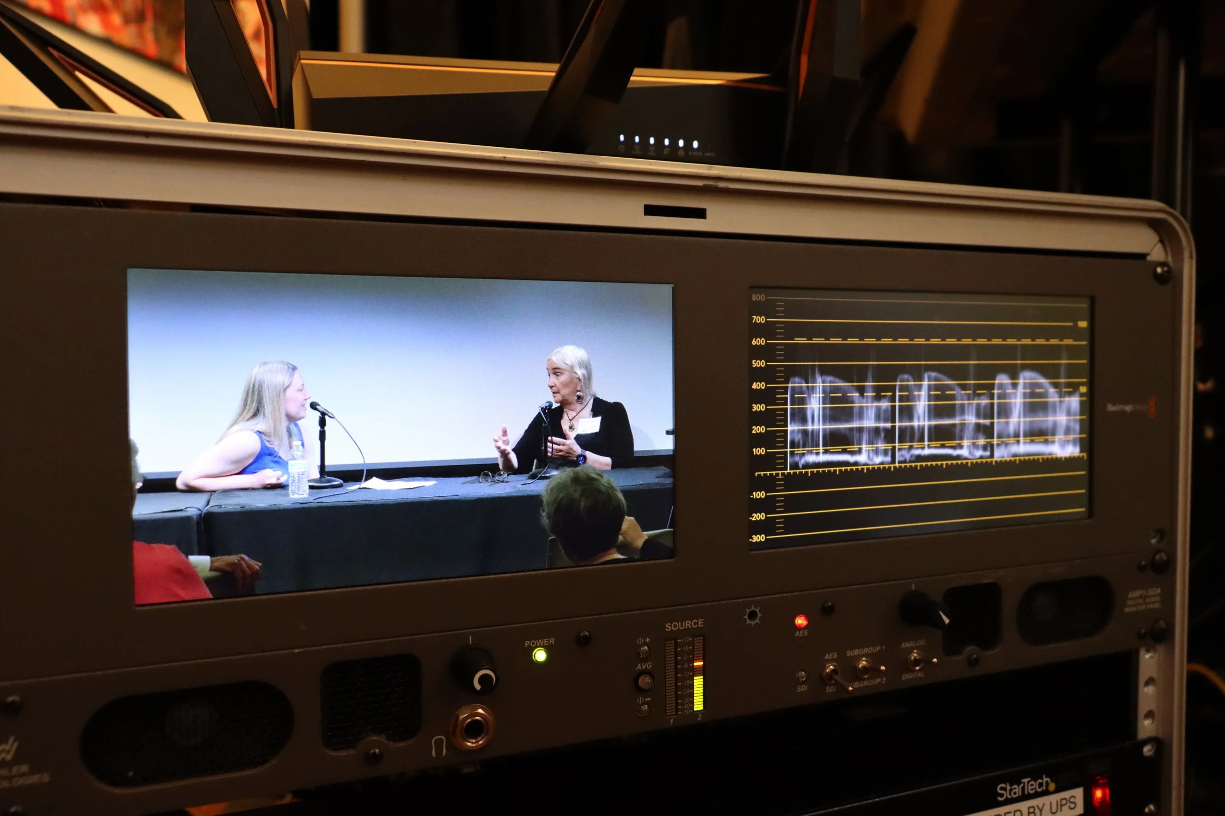 Video monitor showing two women sitting at a table with a microphone, engaged in a discussion during a presentation or interview.