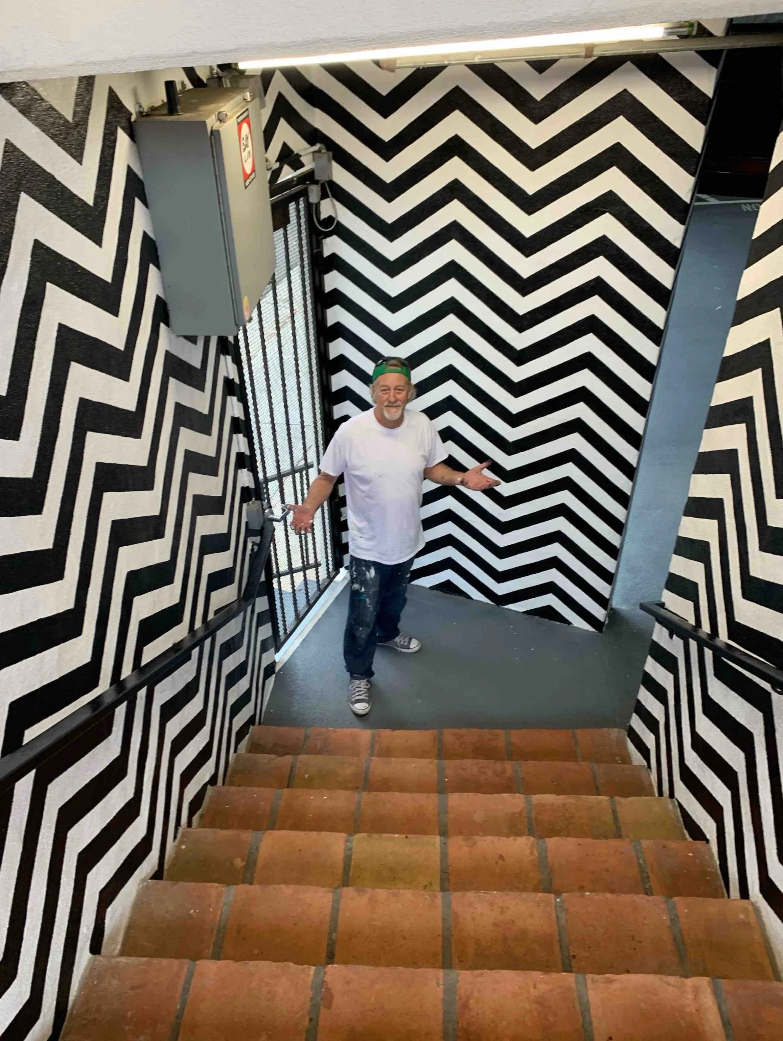 John Glomb standing at the bottom of a staircase with black and white zigzag patterned walls, wearing a white t-shirt, dark jeans, sneakers, and a backwards cap.